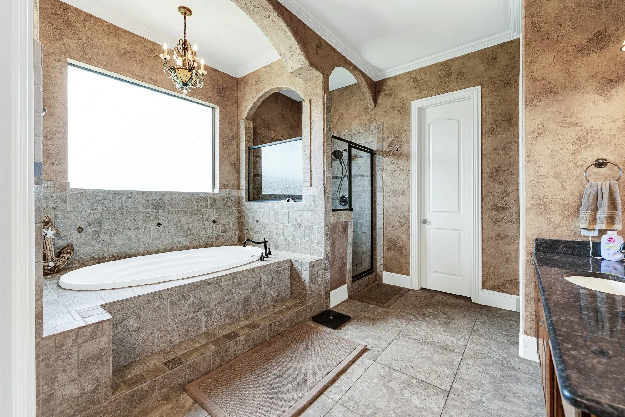 An elegant, tile-encased soaking tub sits beneath a large window, bathing the corner in serene natural light. It is your ultimate evening retreat, where you can sink into a warm bubble bath under the soft glow of the hanging chandelier.