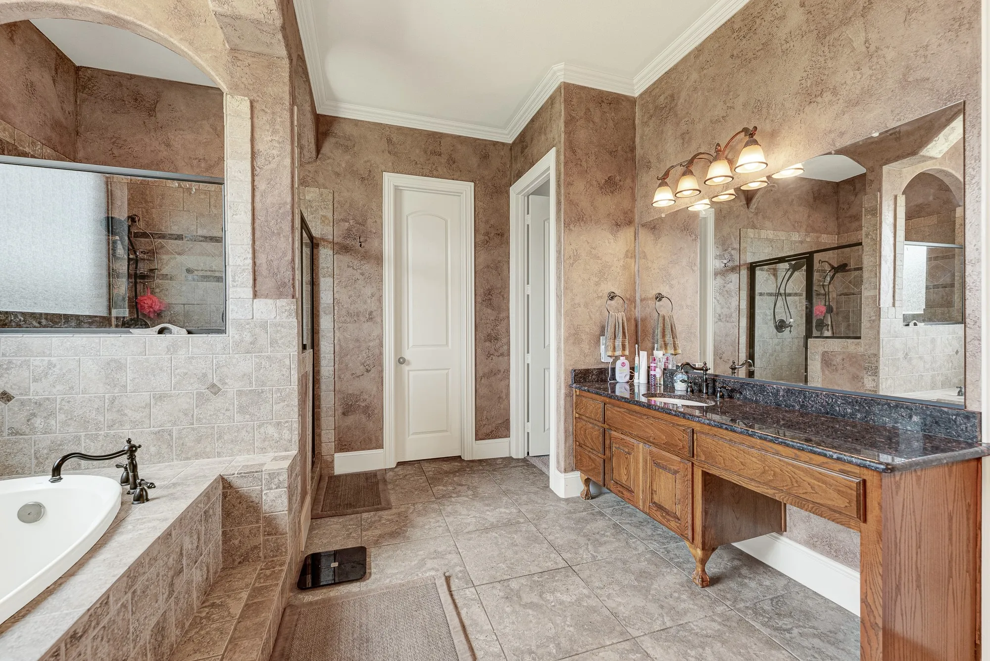 A handsome, wood-crafted vanity with a rich stone countertop pairs beautifully with warm, glowing sconces against the textured walls. Picture yourself taking your time getting ready for an evening out, enjoying the peaceful and organized flow of the space.