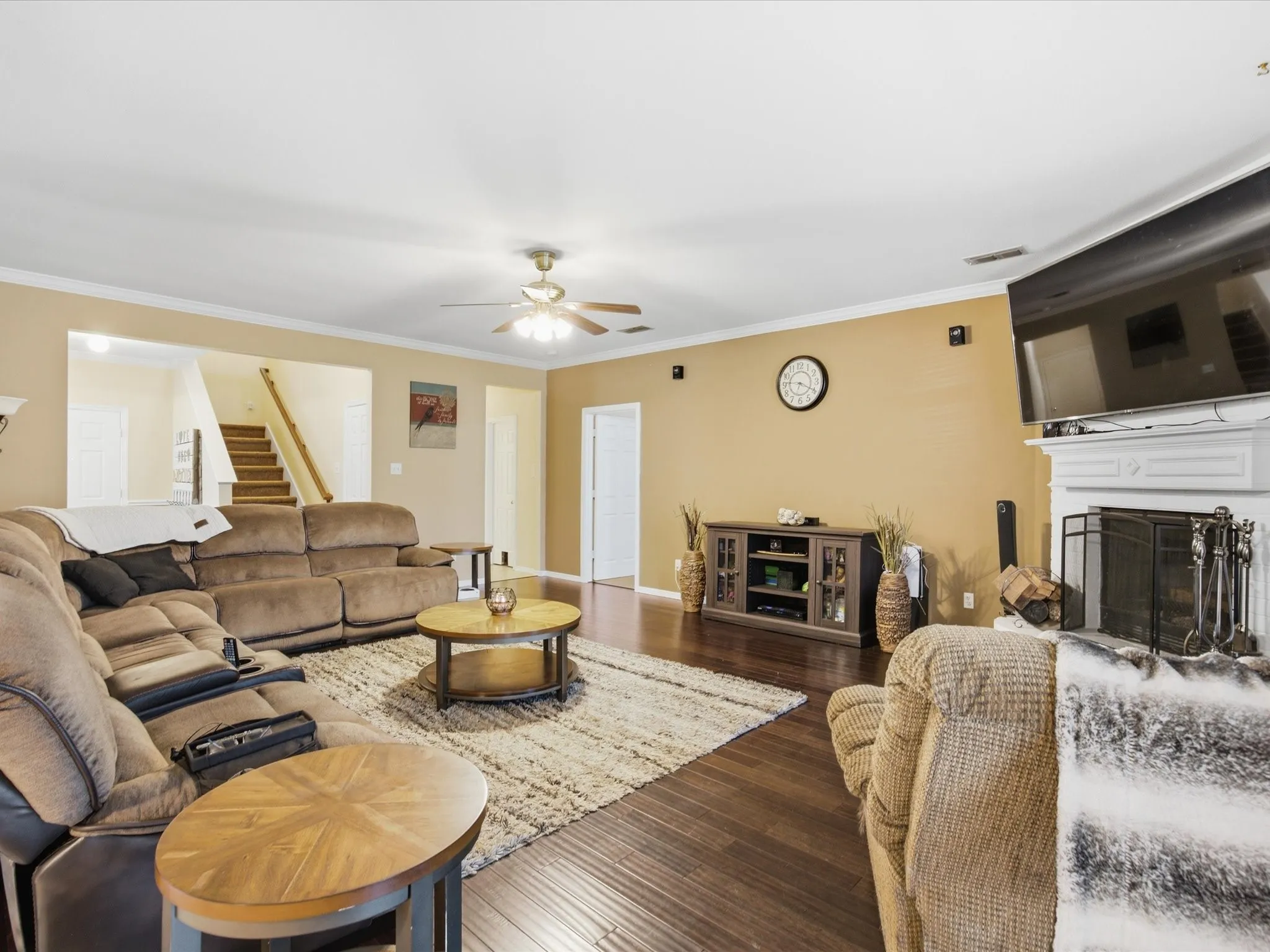 Living area featuring wood-finish flooring, a white mantelpiece, a ceiling fan with integrated lighting, and a visible staircase with wooden handrails