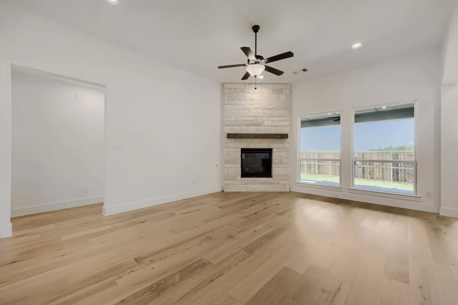 Open-concept living area with light wood-finish flooring, a corner stone-faced fireplace with a wood mantel, and a ceiling fan