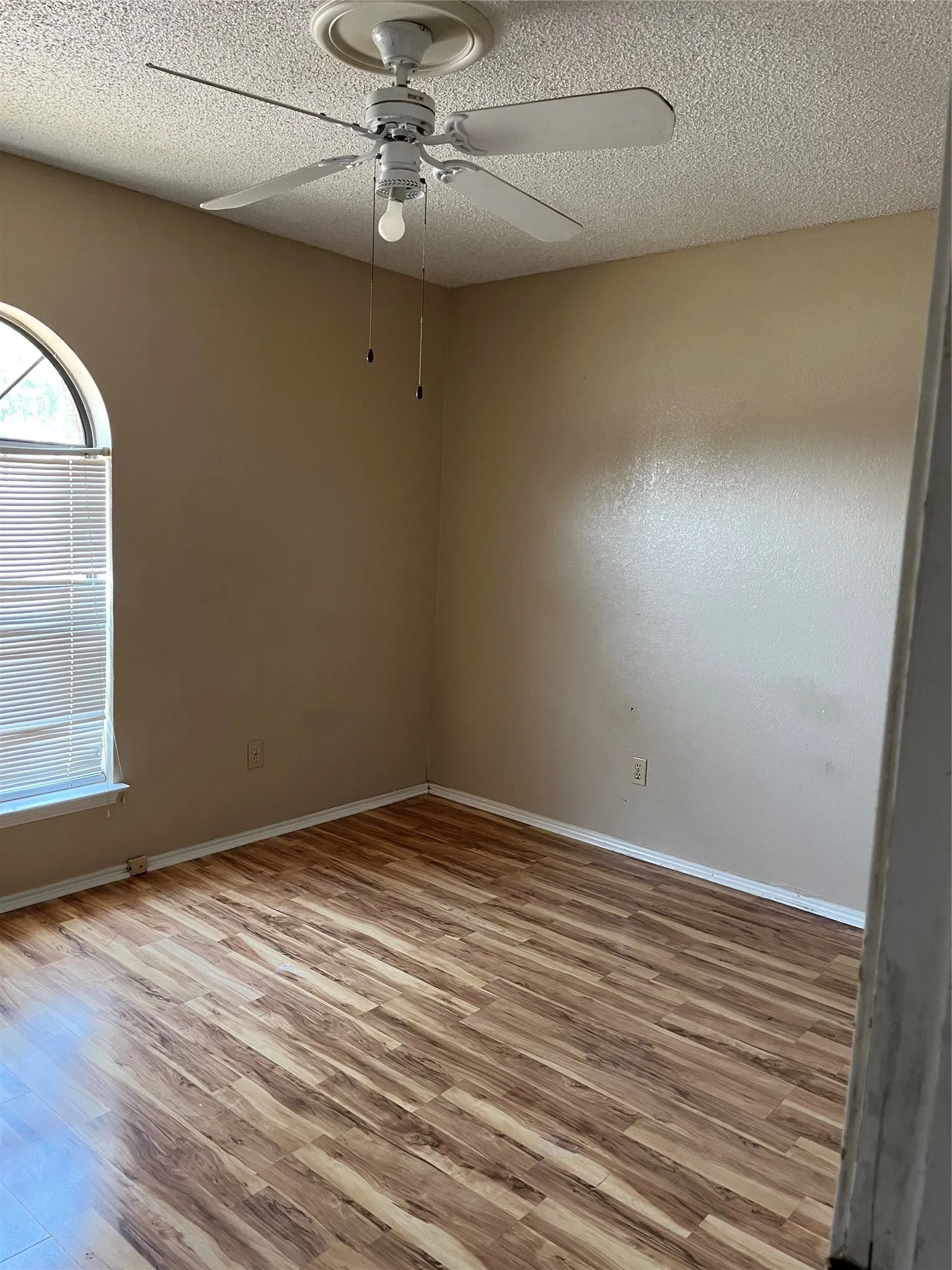 Room featuring wood-finish flooring, a ceiling fan with light fixture, and an arched window with blinds