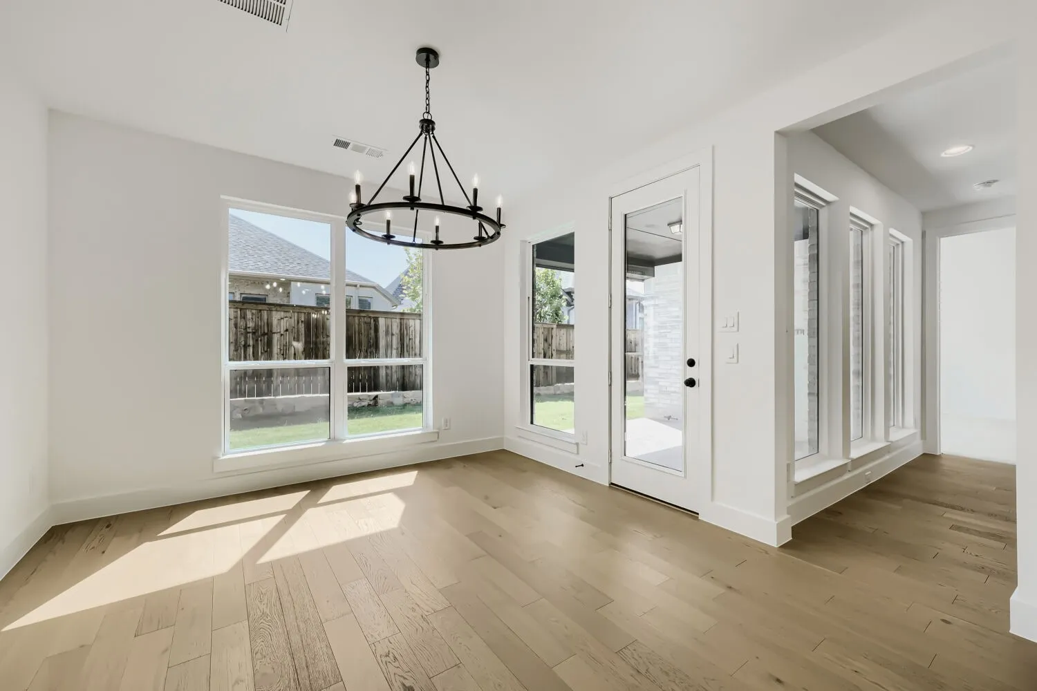 Spacious room featuring light wood-finish flooring, a contemporary black iron chandelier, a large window overlooking the backyard, and a glass-paneled exterior door