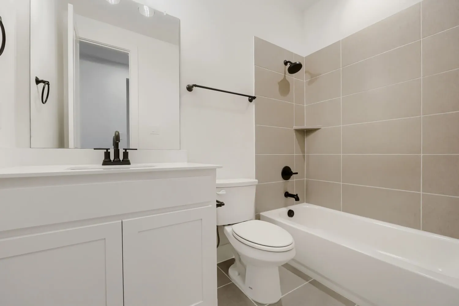 Contemporary bathroom featuring a white vanity with a solid surface countertop, matte black fixtures, a bathtub with a tiled surround, and light-colored floor tiling
