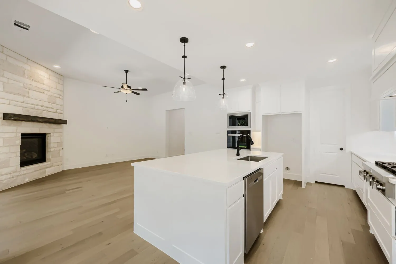 Open-concept living space featuring a stone-clad fireplace with a wood mantel, wood-finish flooring, a contemporary ceiling fan, recessed lighting, and a kitchen island with a sink and stainless steel dishwasher