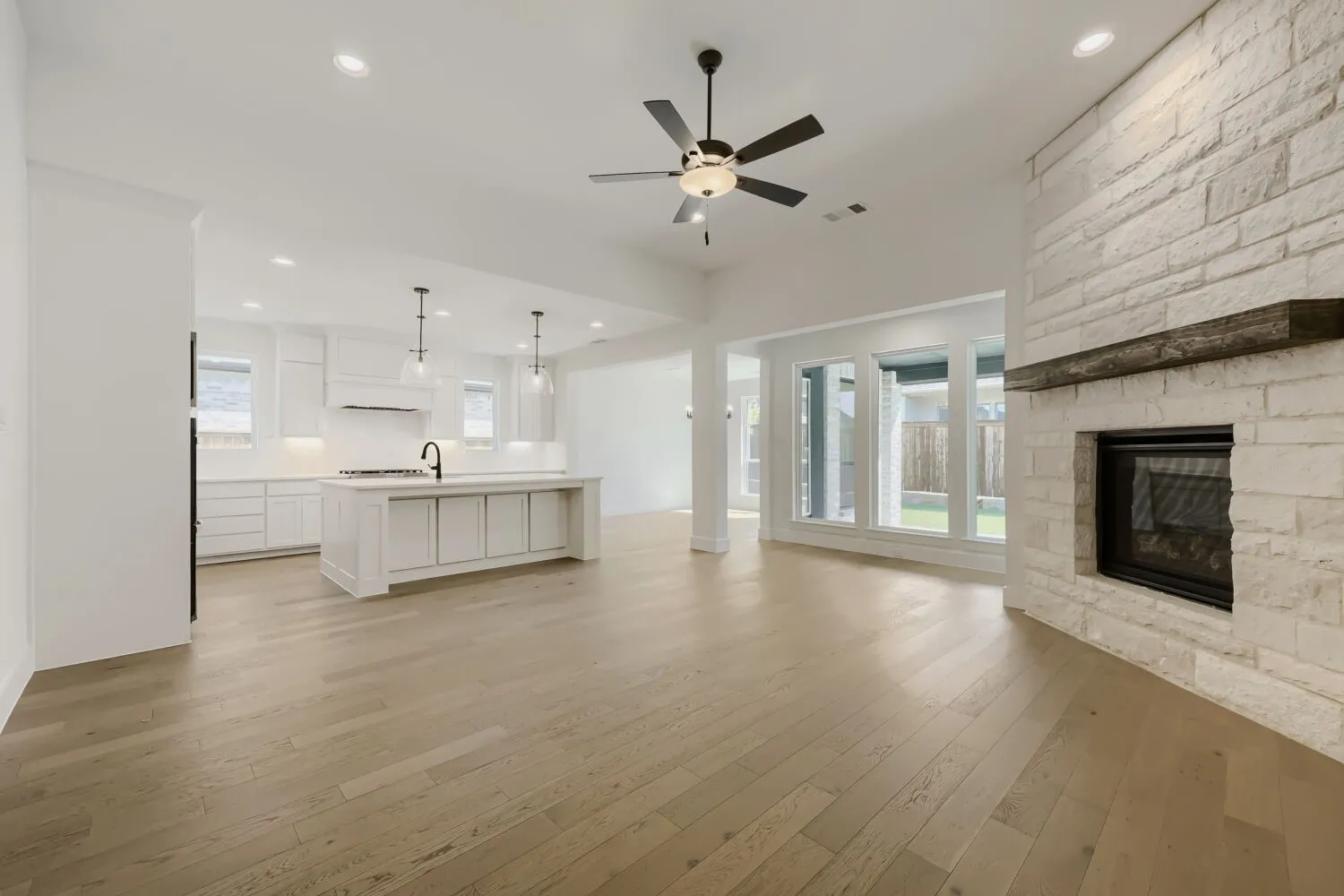 Spacious open-concept living area featuring light wood-finish flooring, a contemporary fireplace with a stone surround and wood mantel, recessed lighting, and a ceiling fan