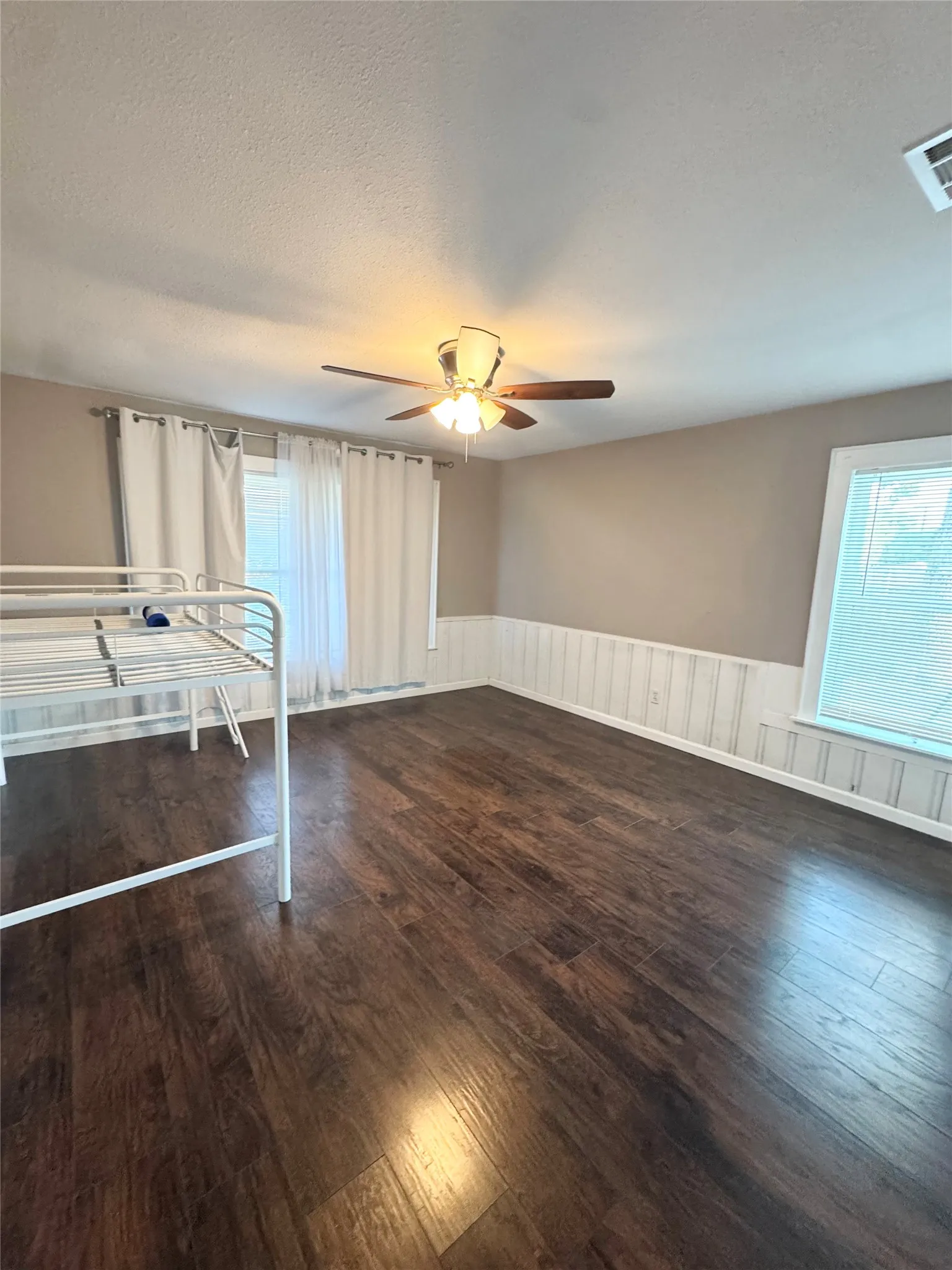 Spacious room featuring wood-finish flooring, a ceiling fan with integrated lighting, two windows for natural light, and wainscoting along the walls