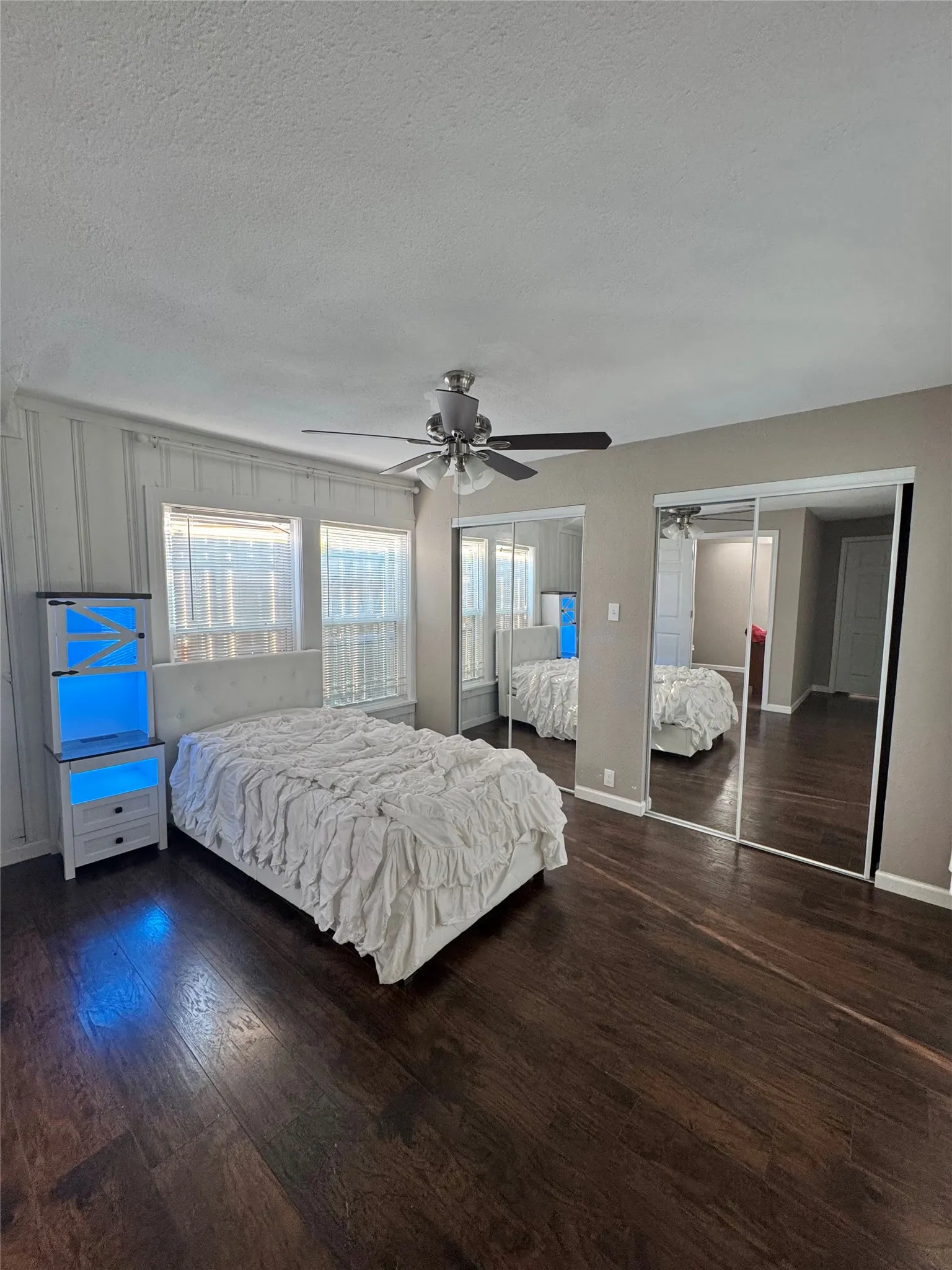 Spacious bedroom featuring dark wood-finish flooring, a ceiling fan with integrated lighting, and multiple windows with blinds
