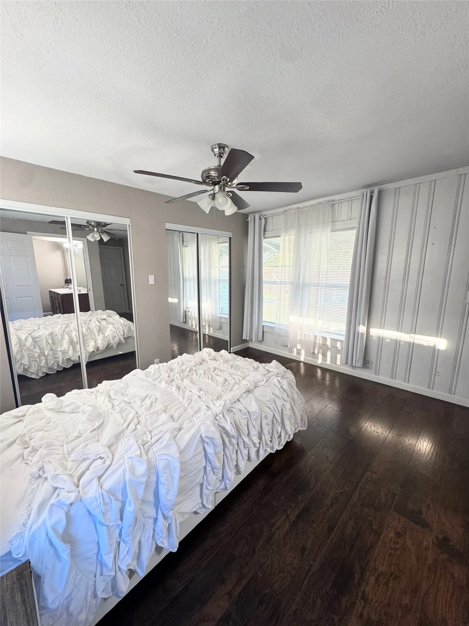 Spacious room featuring dark wood-finish flooring, mirrored closet doors, a ceiling fan with integrated lighting, and multiple windows with sheer and solid curtains