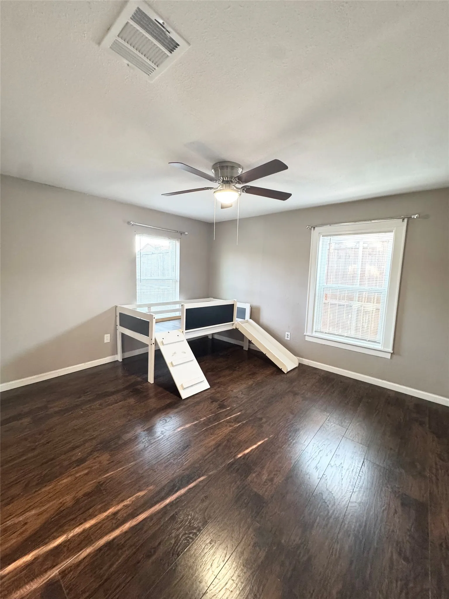 Spacious room featuring rich wood-finish flooring, neutral wall tones, and dual windows