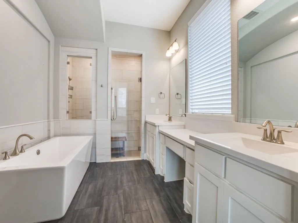Bathroom featuring a freestanding tub, dual vanities with integrated sinks, a glass-enclosed shower, wood-finish flooring, and a large window with blinds
