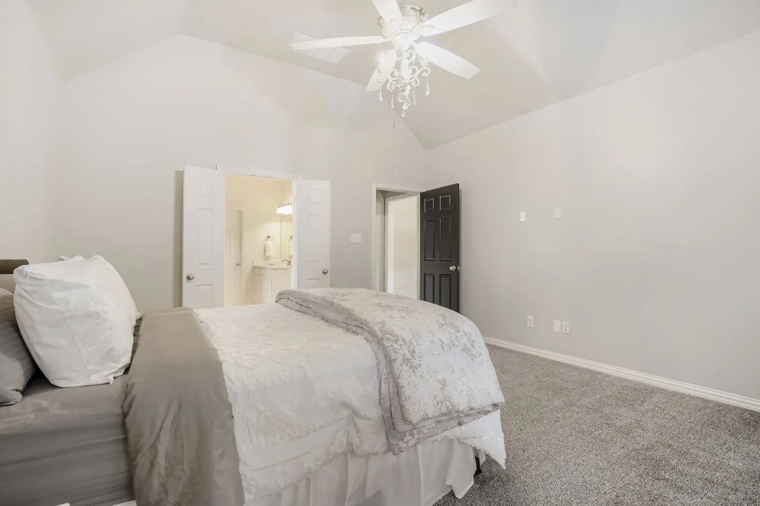 Vaulted ceiling bedroom featuring plush gray carpet, a decorative ceiling fan with chandelier-style lighting, and a dark paneled door
