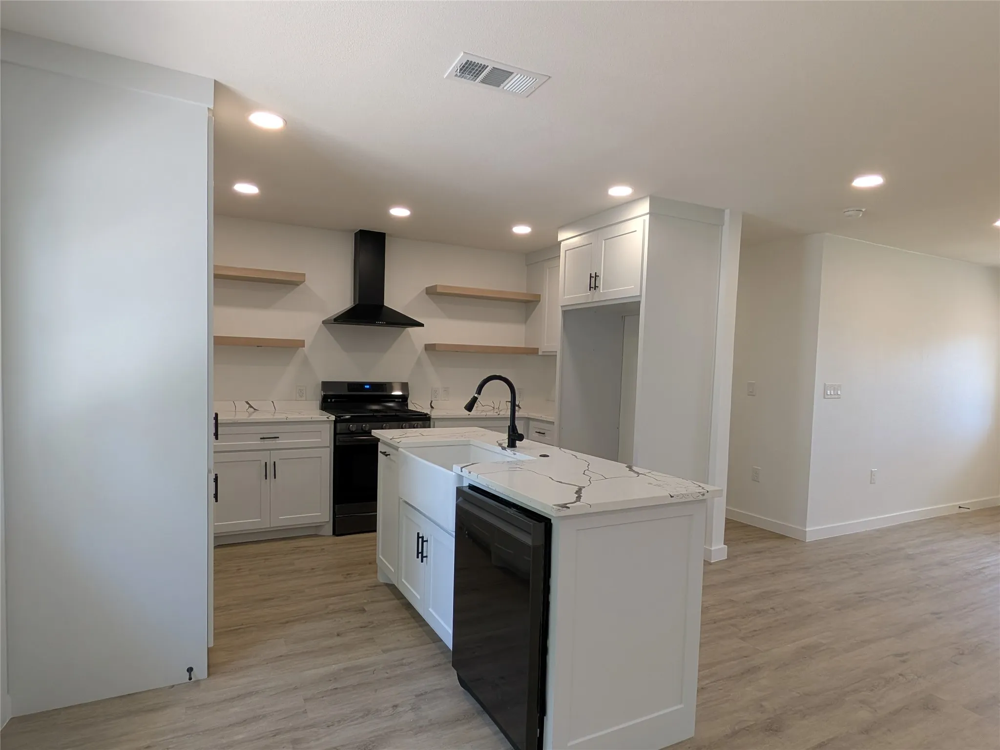 Modern kitchen featuring a central island with an integrated farmhouse sink, white cabinetry with dark hardware, quartz countertops, a black range with a matching range hood, and wood-finish open shelving