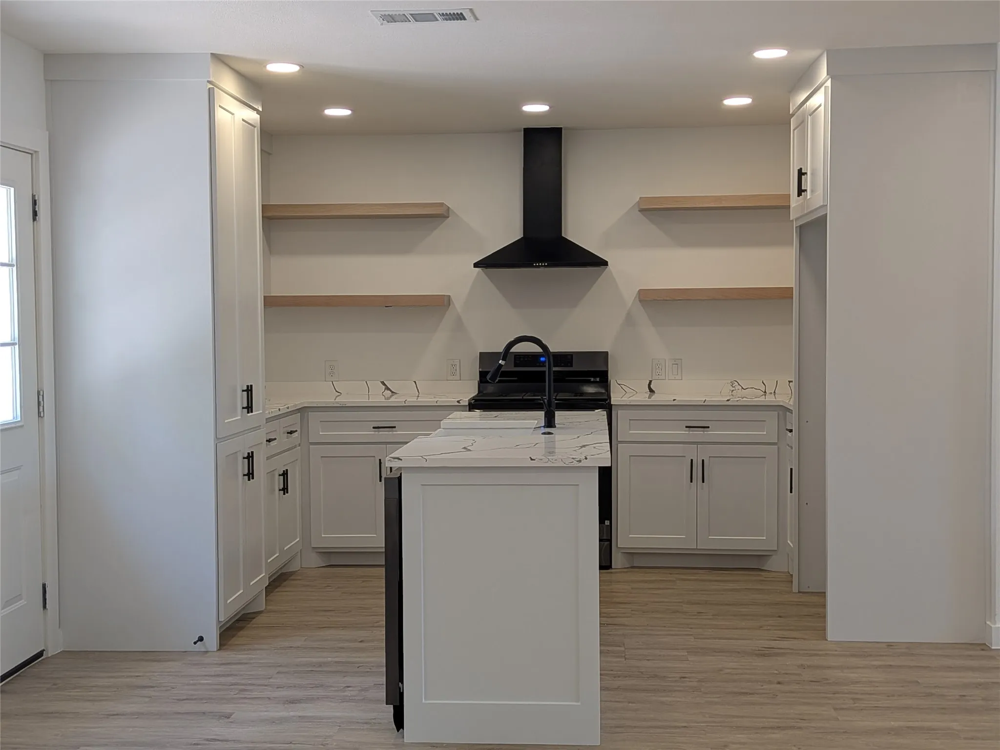 Contemporary kitchen featuring a center island with a built-in sink, white shaker cabinetry, patterned countertops, a black range hood, and wood-finish open shelving