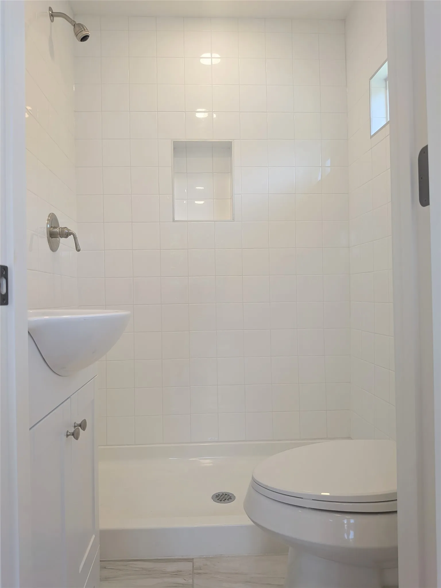 Compact bathroom featuring white square tile walls, a built-in shower niche, and a wall-mounted shower head with a brushed nickel finish