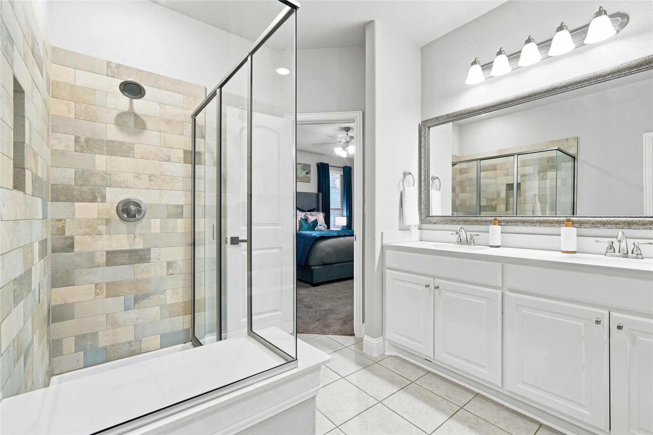Contemporary bathroom featuring a spacious walk-in shower with varied rectangular tiling, a dual-sink vanity with white cabinetry, a large framed mirror, and tile flooring