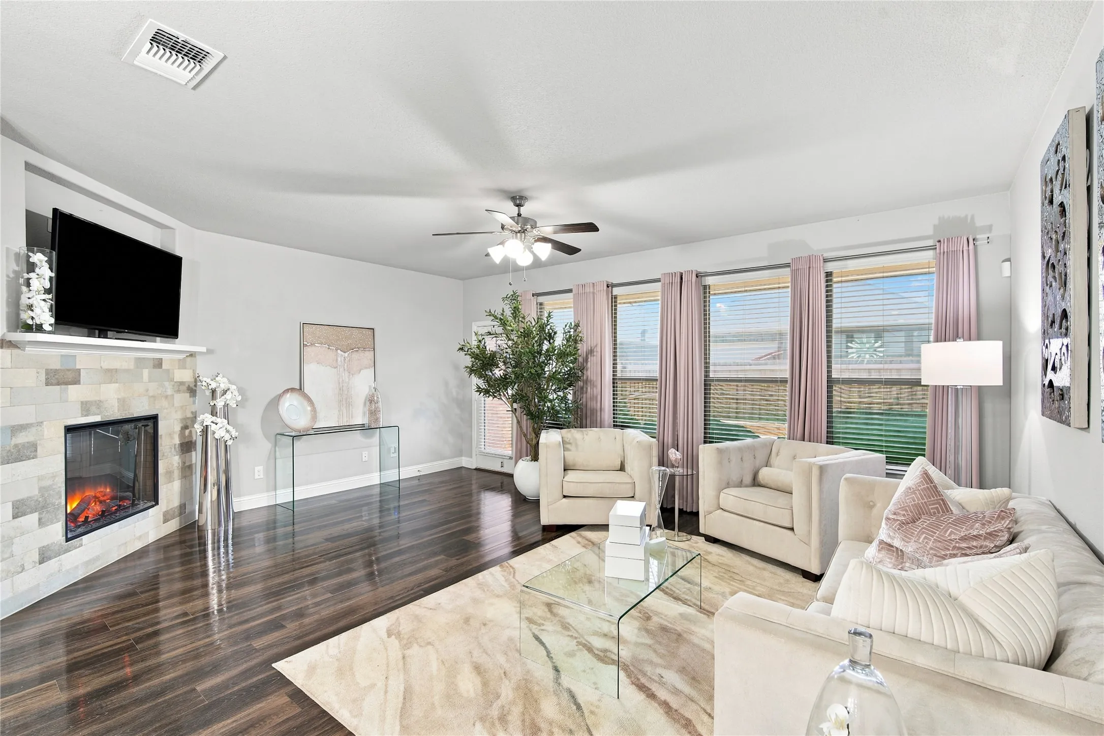 Living area featuring dark wood-finish flooring, a contemporary fireplace with a tile surround and mantle, a ceiling fan with integrated lighting, and multiple windows with blinds