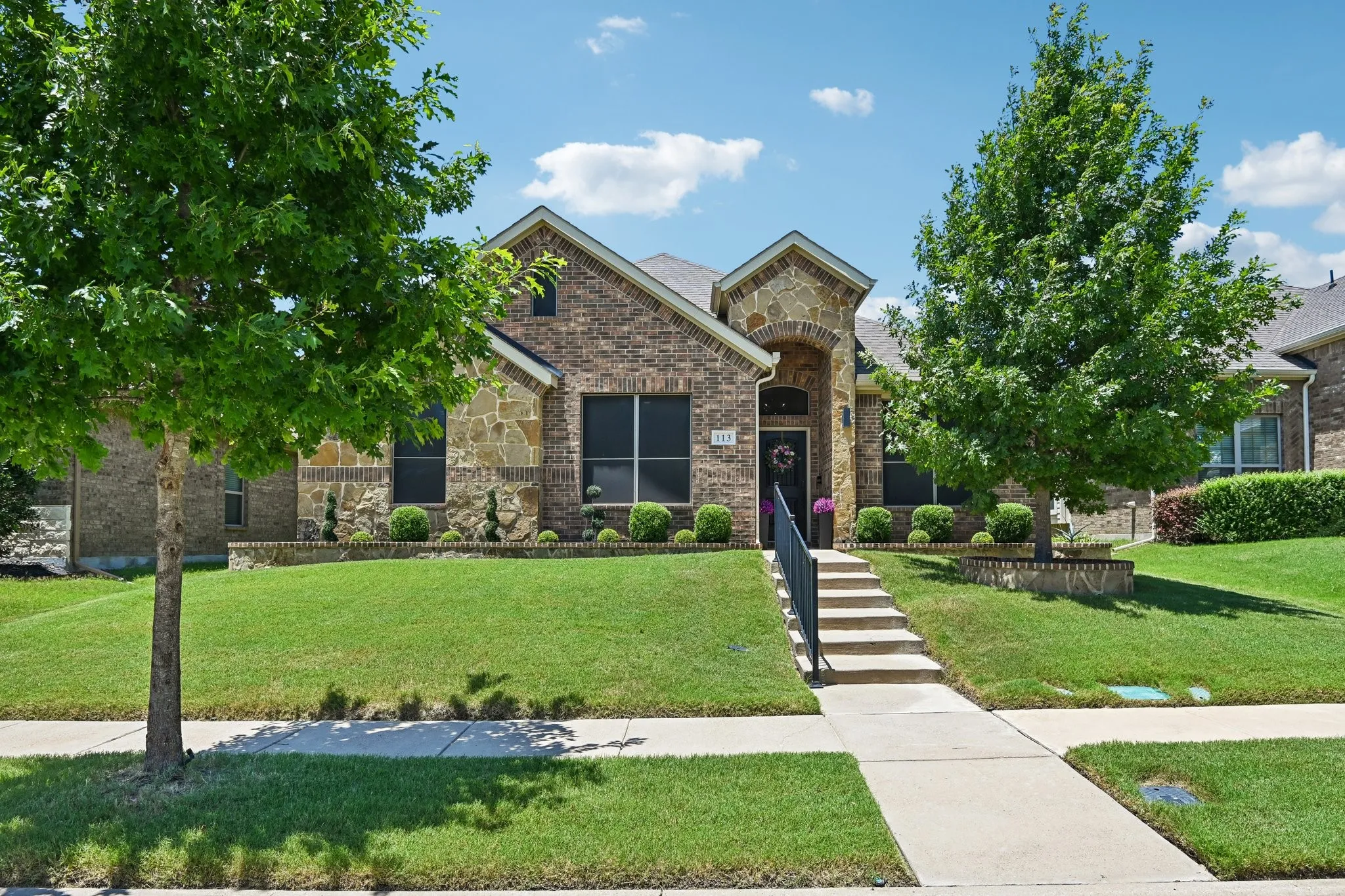 Brick and stone facade residence featuring a covered entry, tiered landscaping, mature trees, and a concrete pathway leading to the front door