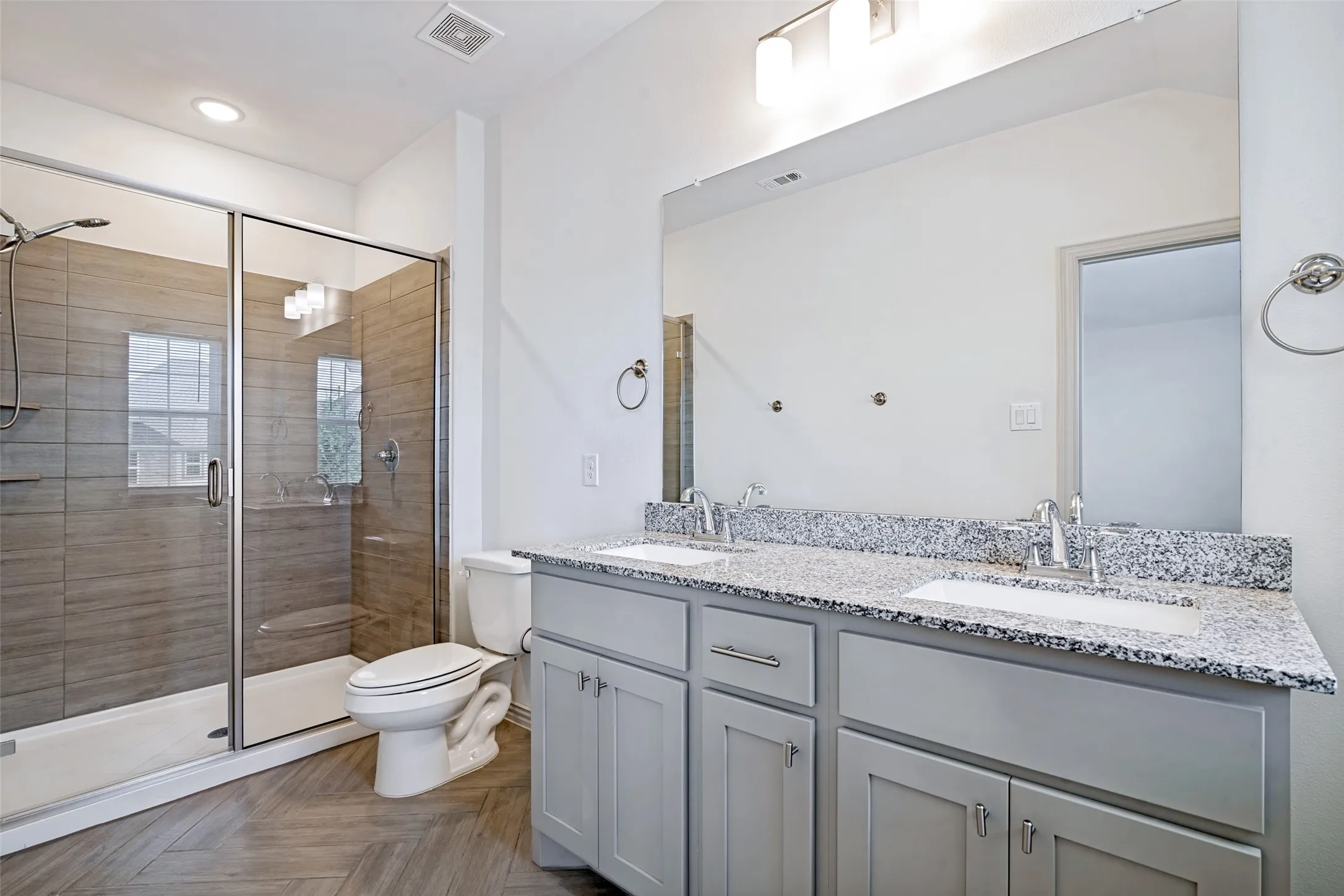 Contemporary bathroom featuring a spacious glass-enclosed shower with horizontal tile work, a dual vanity with light gray cabinetry, and a speckled countertop