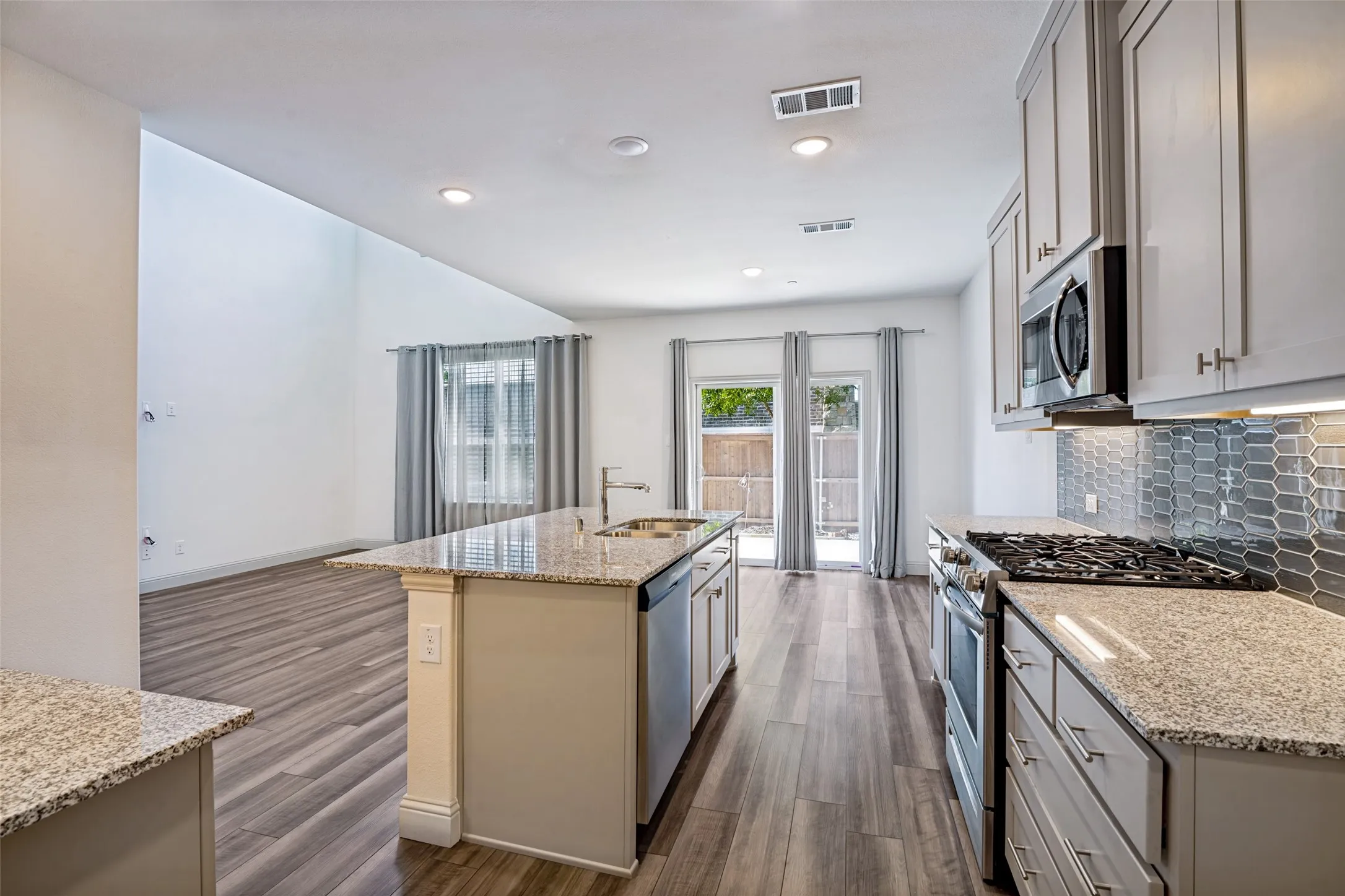 Kitchen with wood-finish flooring featuring granite countertops, stainless steel appliances, a tile backsplash, and an island with a built-in sink