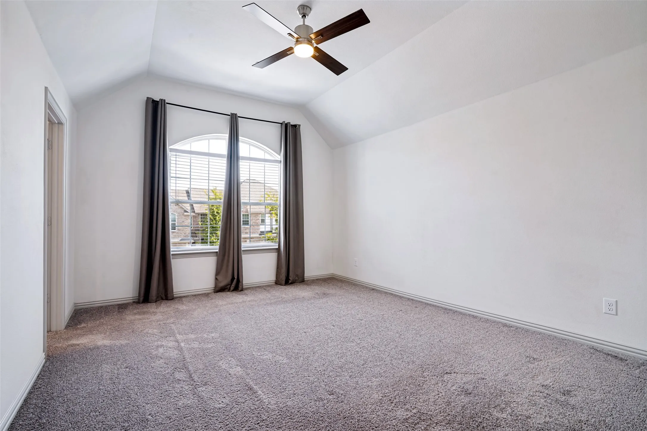 Spacious room featuring vaulted ceilings, a large arched window with blinds, a ceiling fan, and carpeted flooring
