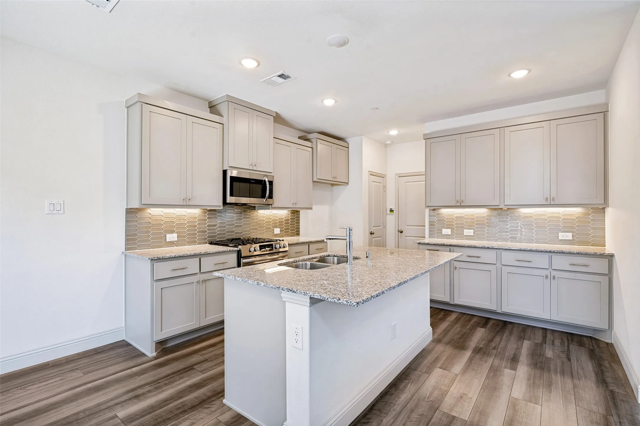 Open-concept kitchen featuring an island with a double sink, light grey cabinetry, stainless steel appliances, granite countertops, and wood-finish flooring