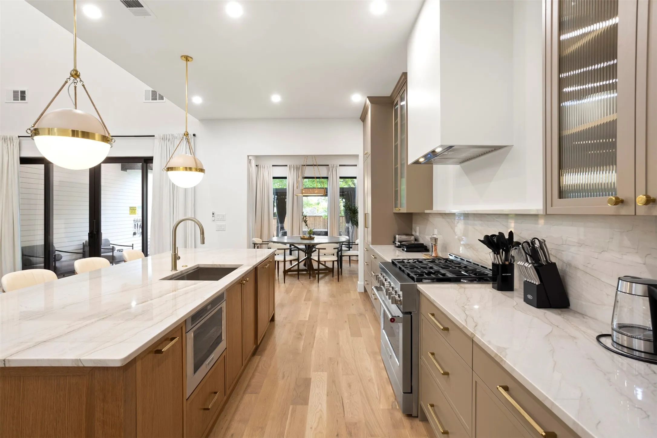 Kitchen featuring light wood-finish flooring, a large central island with an integrated sink and microwave, pendant lighting, stone countertops with a matching backsplash, and stainless steel appliances