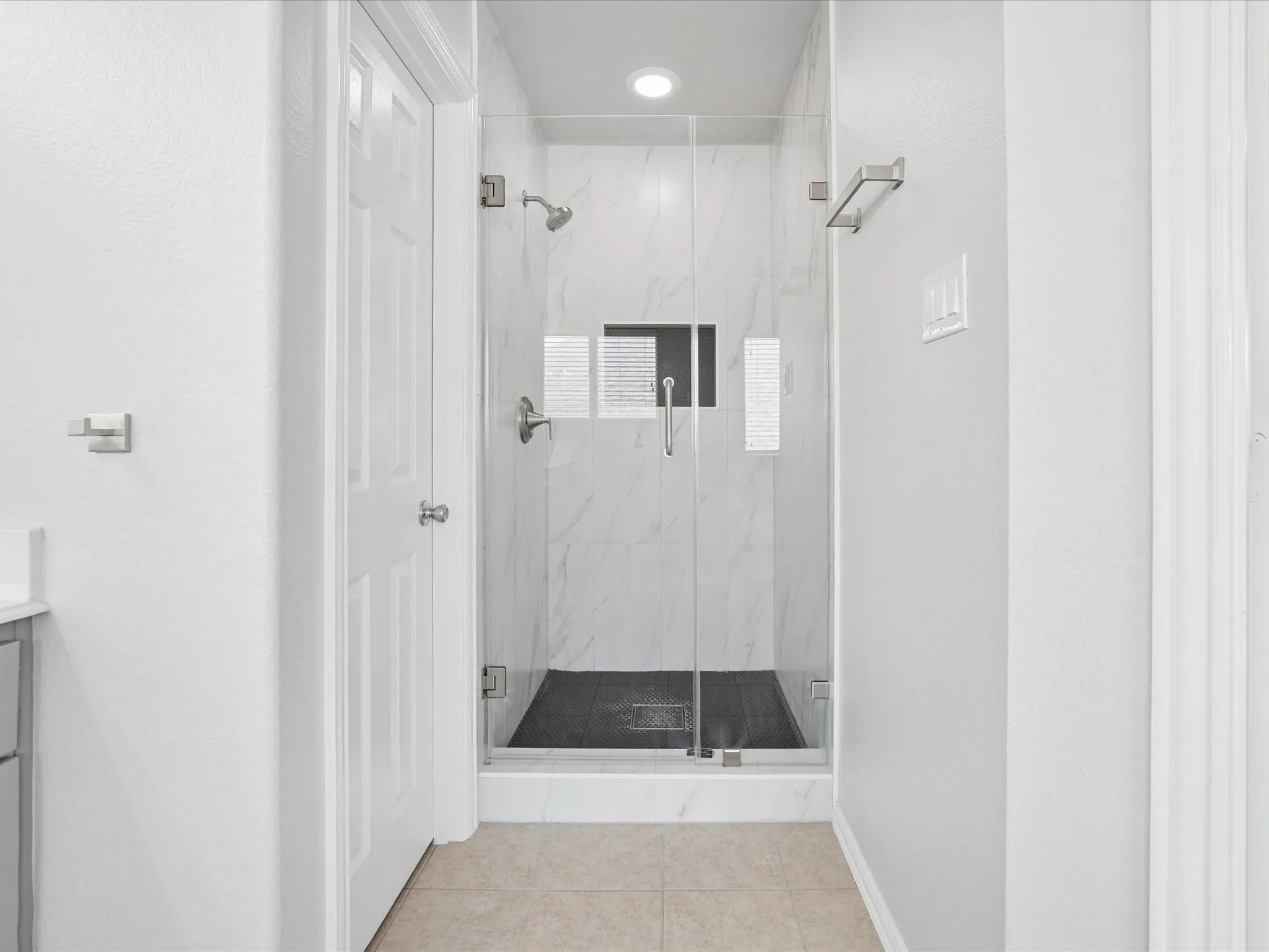 Glass-enclosed shower featuring white-veined stone tile, a built-in shower niche, and a dark tile shower floor