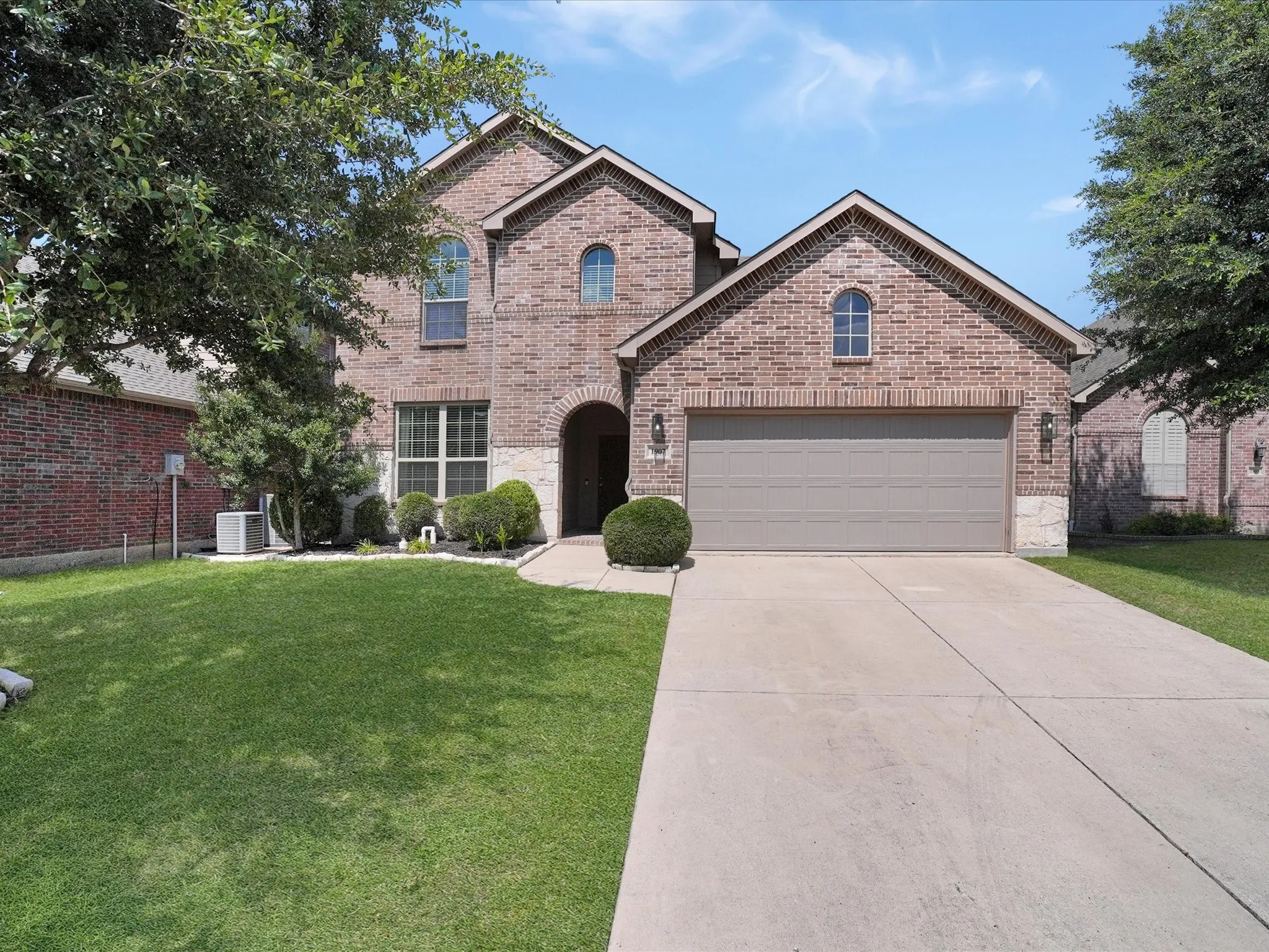 Brick and stone facade featuring a two-car garage, concrete driveway, arched entryway, and mature landscaping