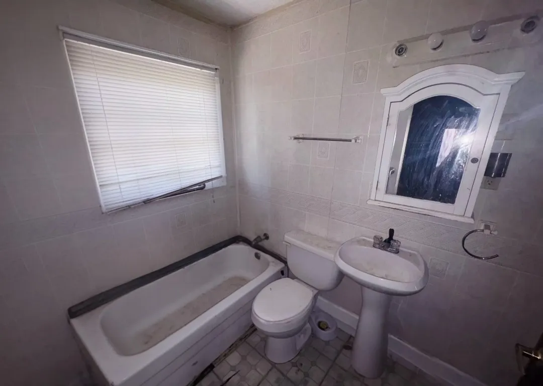 White tiled bathroom featuring a single window with blinds, a pedestal sink, a toilet, and a bathtub