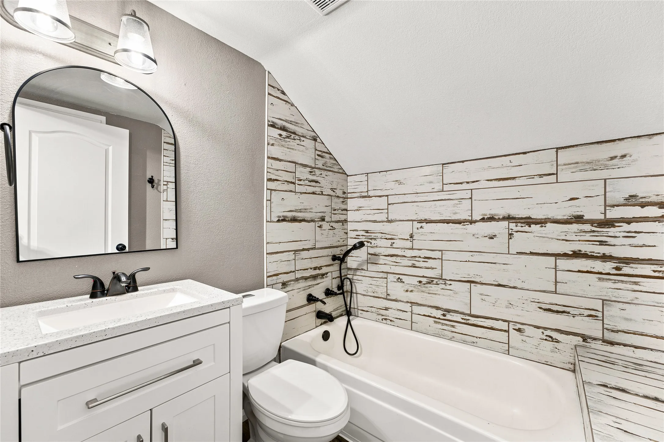 Bathroom featuring a white vanity with a light-colored countertop, an arched mirror, white subway tile-style wall paneling, a white bathtub, and contemporary fixtures