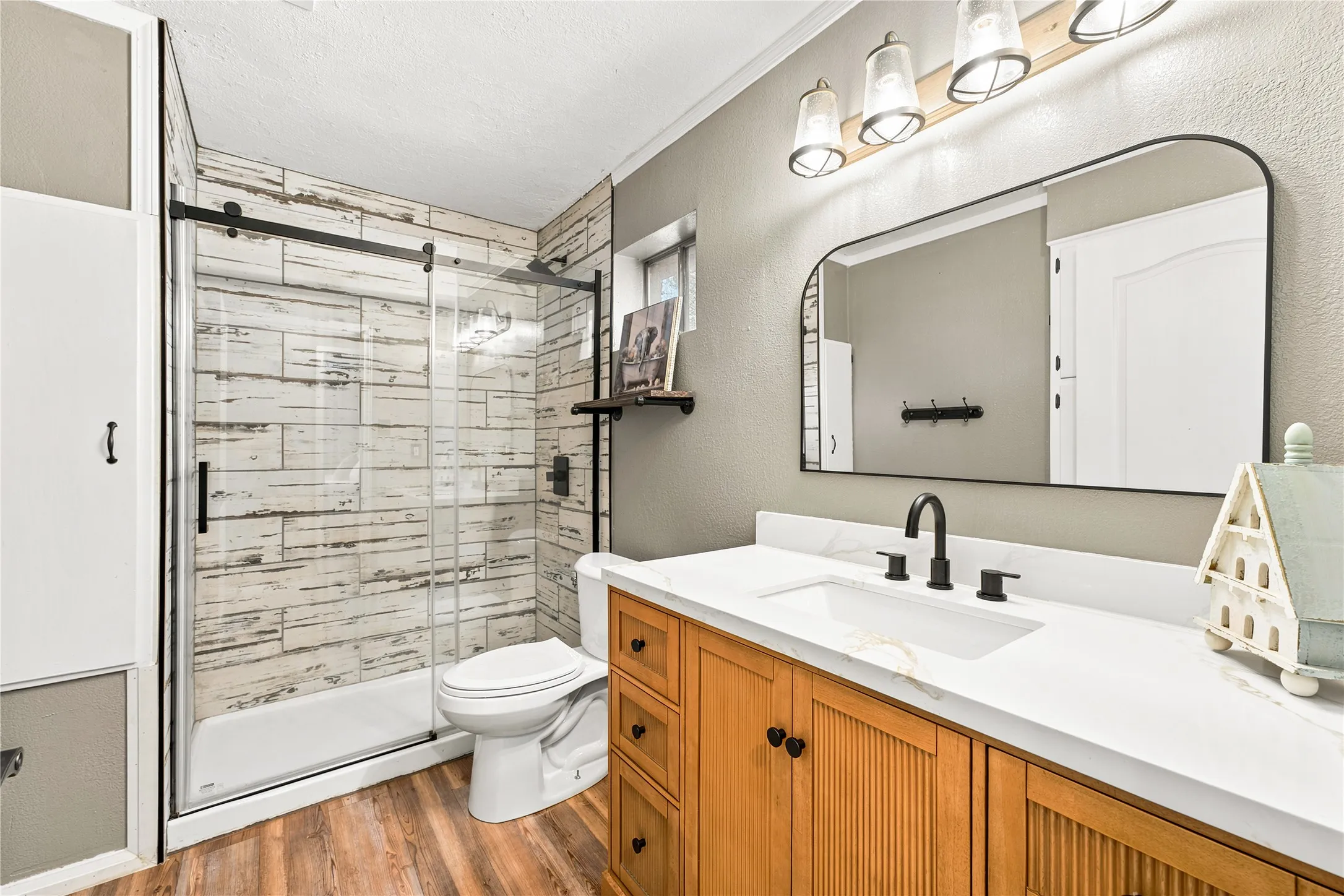 Bathroom featuring wood-finish flooring, a wood vanity with a white countertop and undermount sink, a large arched mirror, and a walk-in shower with a sliding glass door and white-washed wood-look tile surround