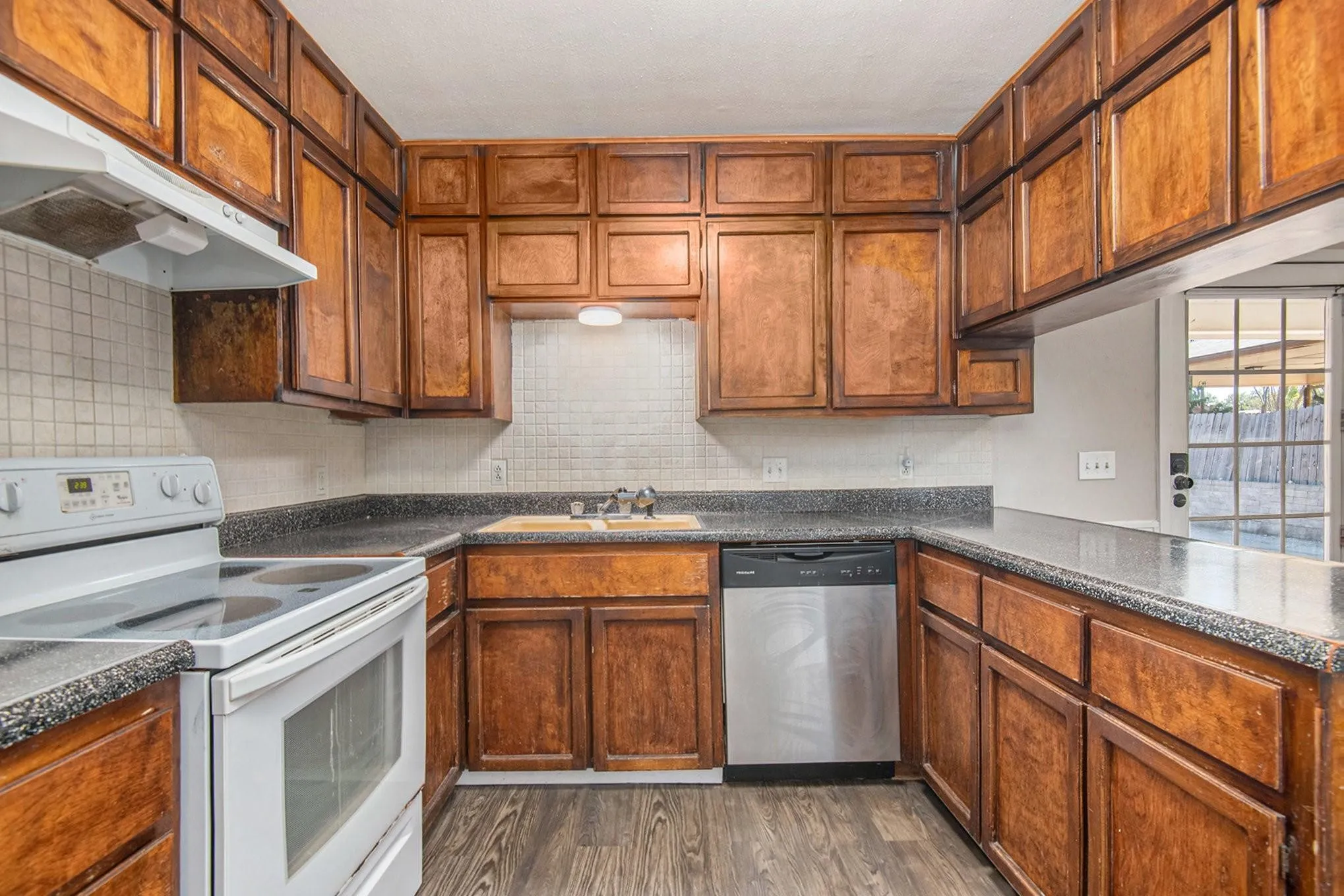 Kitchen featuring wood cabinetry, speckled countertops, a white tile backsplash, wood-finish flooring, and a stainless steel dishwasher