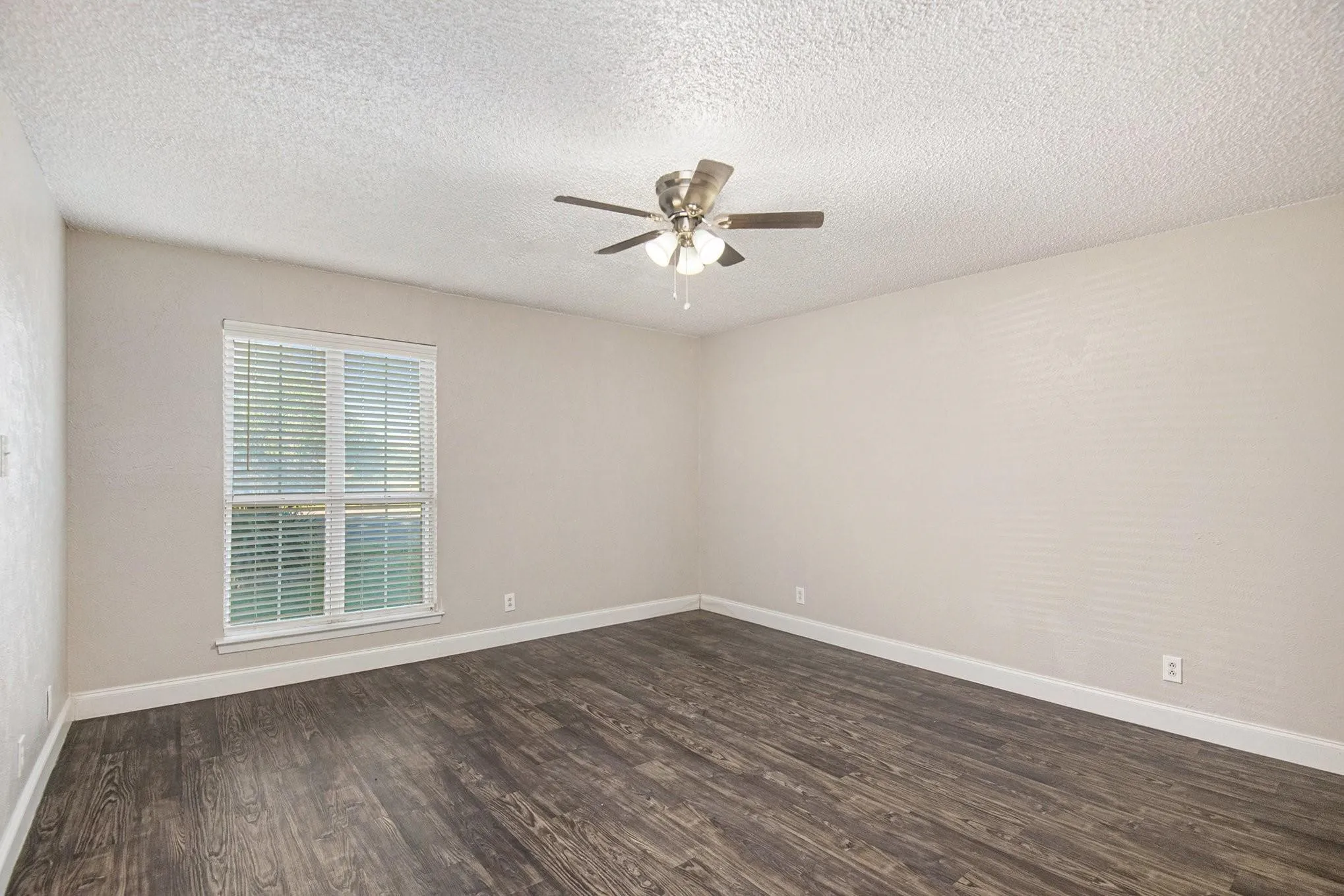 Spacious room featuring dark wood-finish flooring, light-colored walls, a ceiling fan with integrated lighting, and a large window with blinds