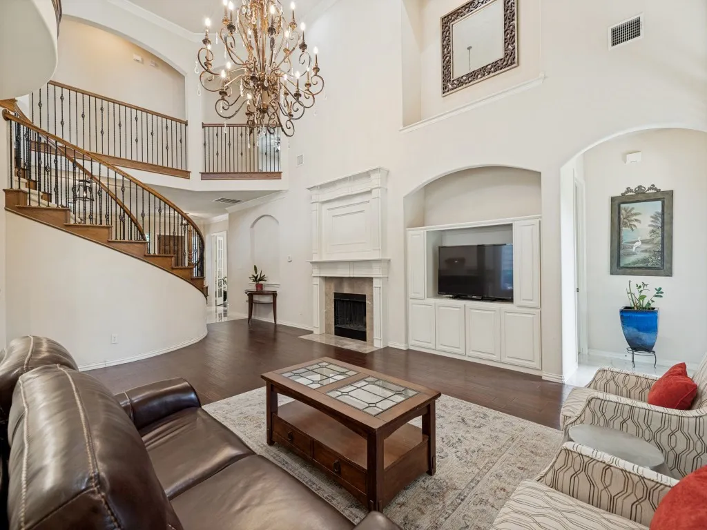 Two-story living area featuring a grand curved staircase with wrought iron balusters, a stone fireplace with a substantial mantel, dark wood-finish flooring, and built-in media cabinetry