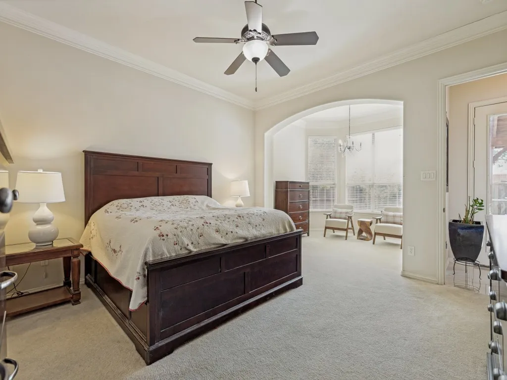 Carpeted bedroom featuring a ceiling fan, crown molding, and an arched entryway leading to a bay window alcove with a chandelier and outdoor access