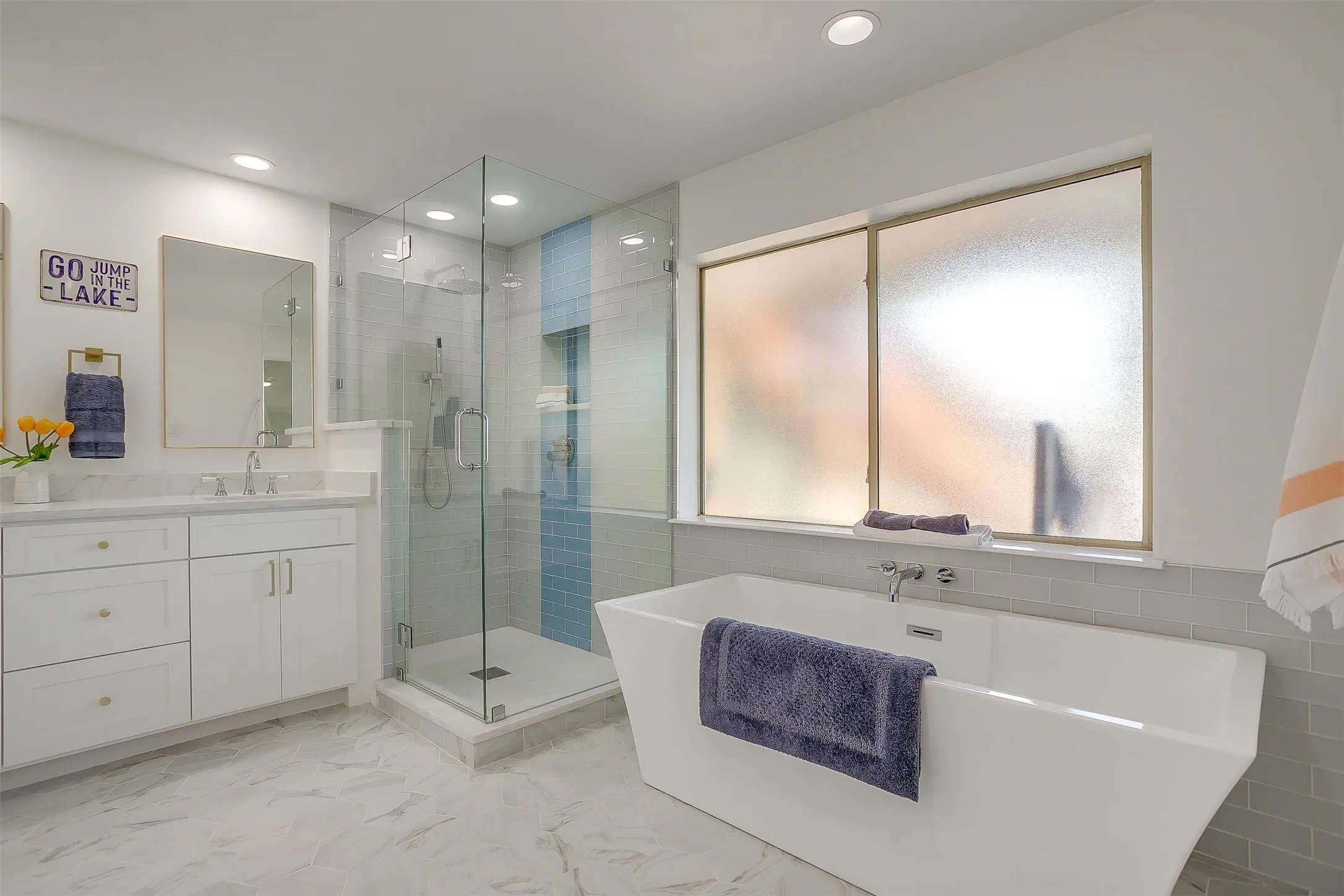 Bathroom featuring a freestanding soaking tub, a glass-enclosed shower with blue and white tile accents, a double vanity with white cabinetry and a white countertop, recessed lighting, and large format floor tiling