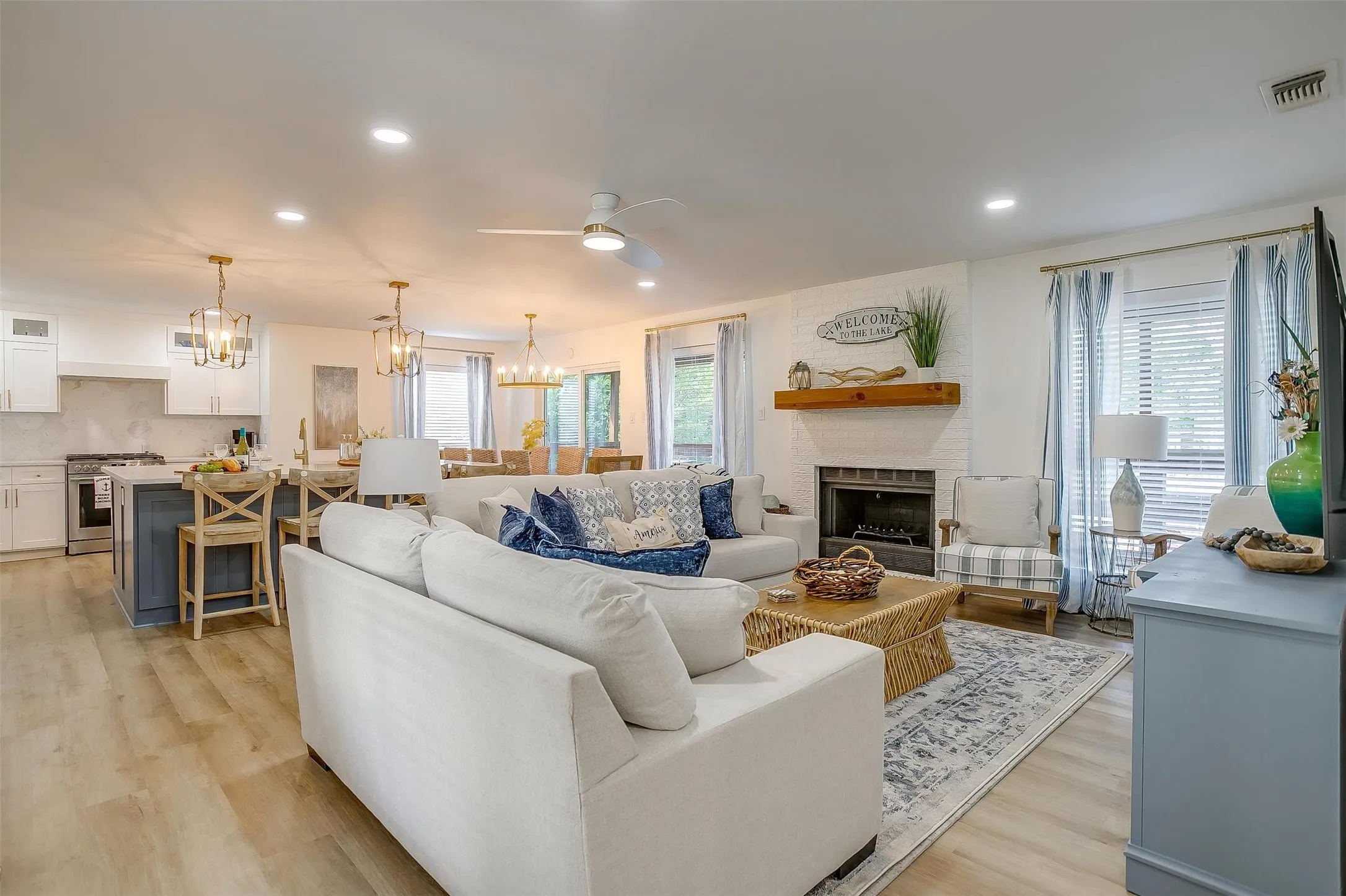 Open-concept living space featuring wood-finish flooring, a white brick fireplace with a wood mantel, and multiple large windows