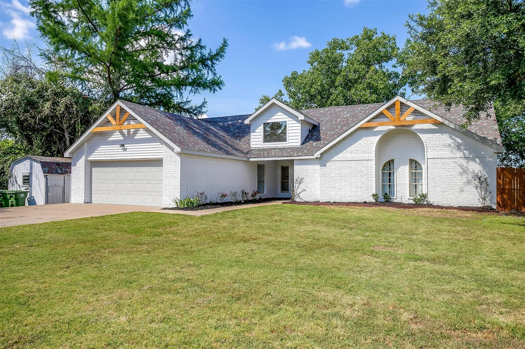 White brick and siding exterior with wood-beam gable accents, arched window alcove, two-car garage, and a separate storage shed