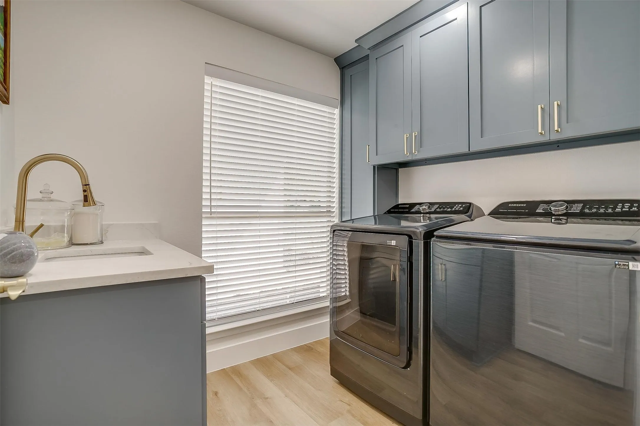 Laundry area featuring modern blue cabinetry, a built-in sink with a gold-tone faucet, a large window with blinds, light wood-finish flooring, and white walls