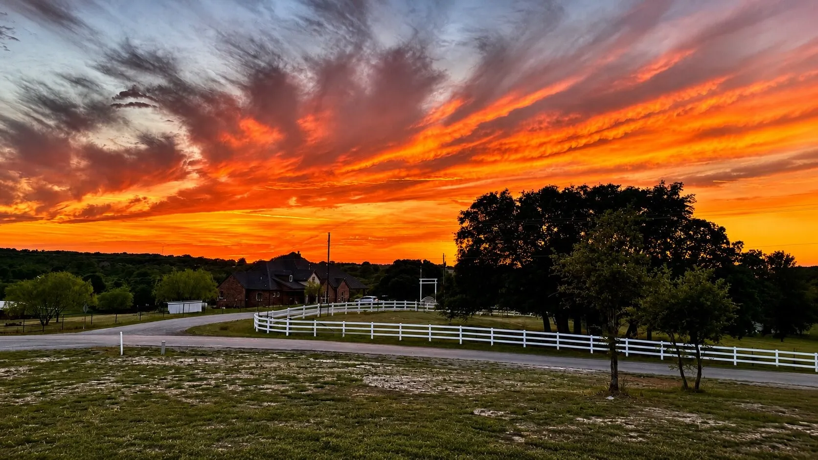 Expansive rural landscape featuring a single-story stone exterior residence, a paved driveway, a white fencing system, and mature trees