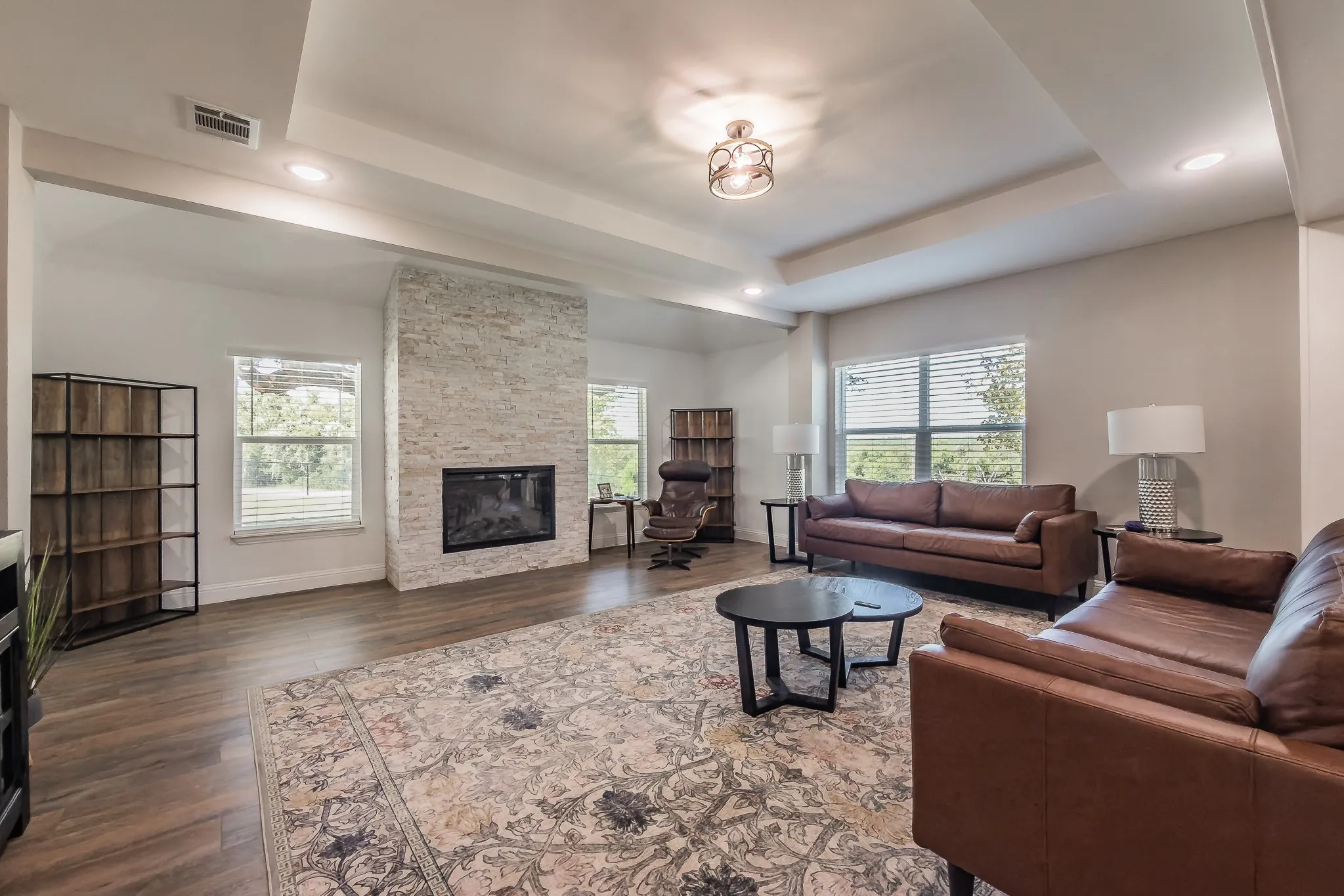 Spacious living area featuring wood-finish flooring, a stacked stone fireplace with a black insert, and a tray ceiling with recessed lighting