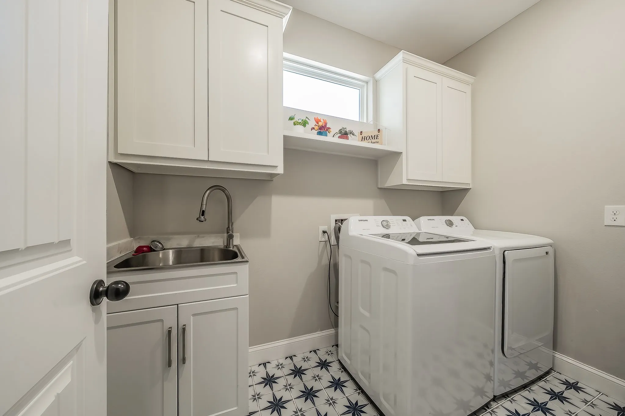 Laundry room featuring patterned tile flooring, white shaker-style cabinetry, an under-mount stainless steel sink with a gooseneck faucet, and a high transom window