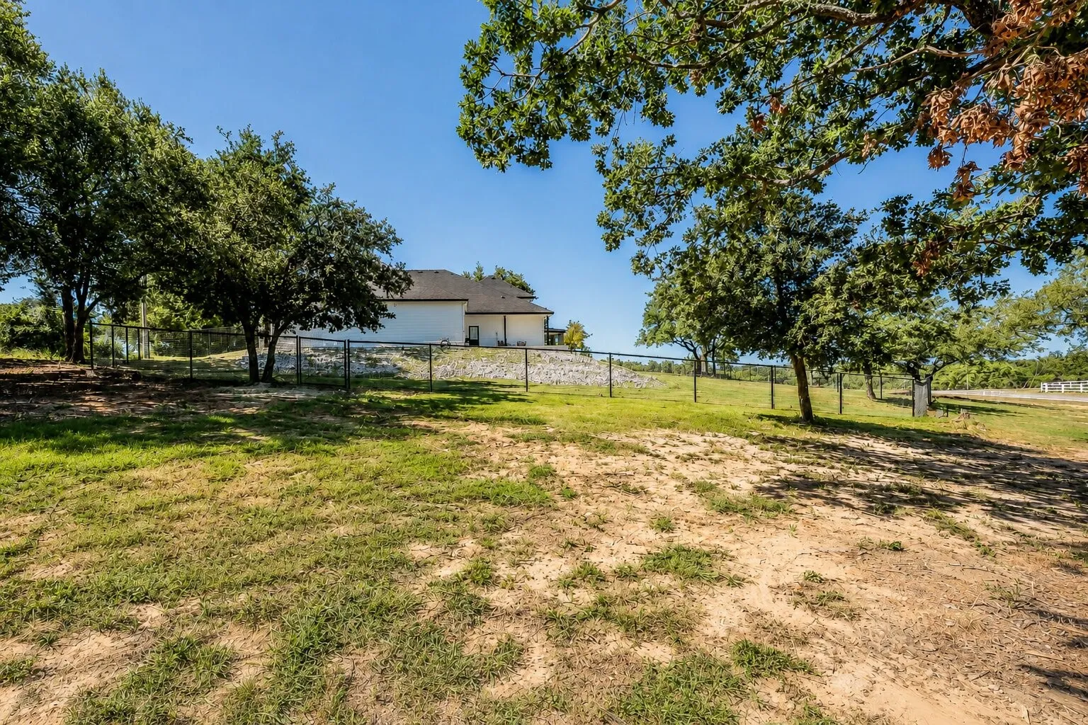Expansive outdoor space featuring a partial chain-link fence, mature trees, and a distant property structure