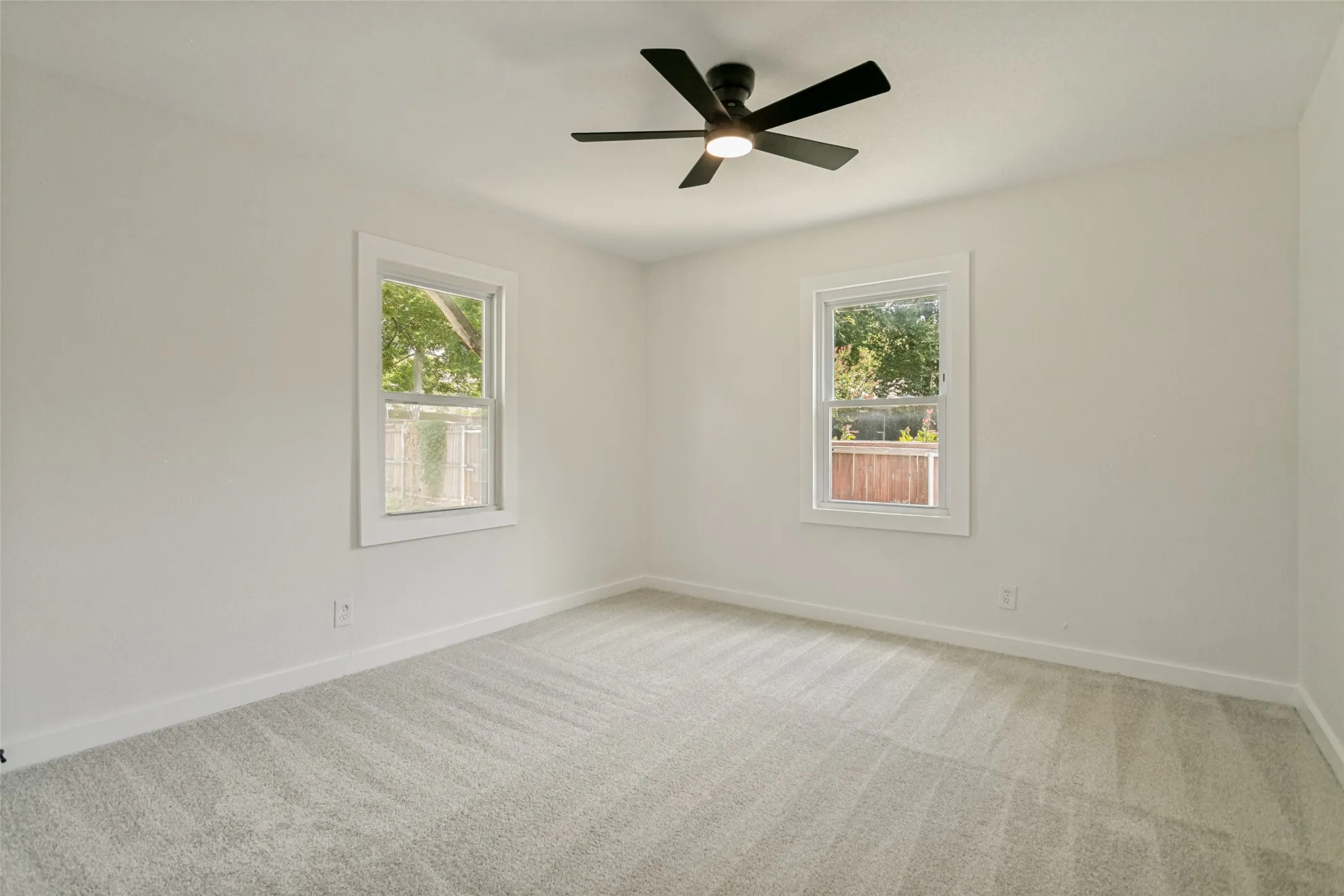 Room featuring light gray carpeting, neutral wall finishes, white trim, two double-hung windows, and a black ceiling fan with integrated lighting