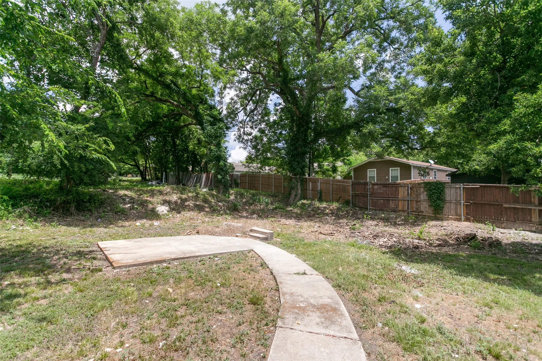 Concrete walkway leading to a concrete patio, surrounded by mature trees and a wood privacy fence
