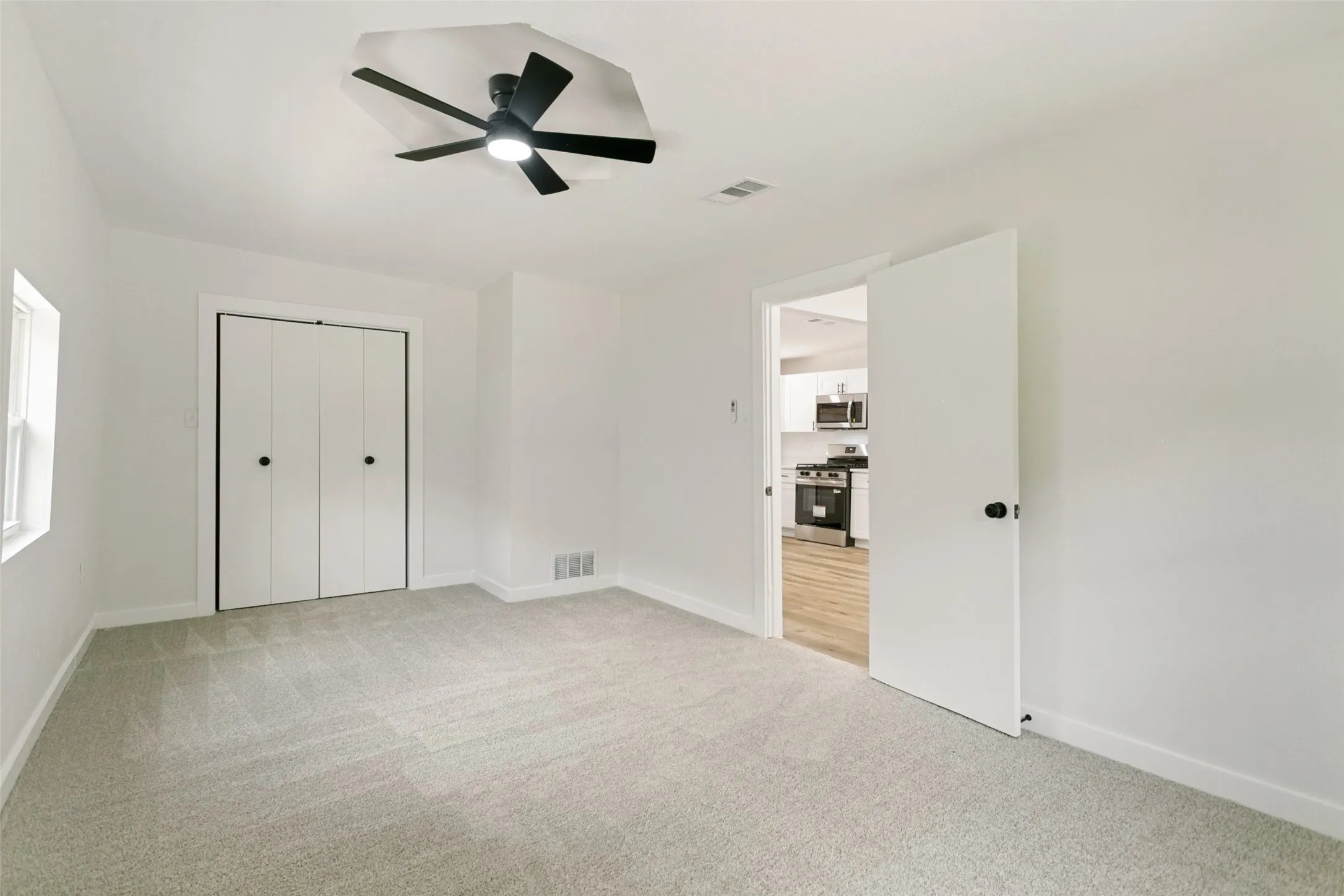 Spacious room featuring light-colored carpeting, a white bi-fold closet, a window providing natural light, a ceiling fan with black blades, and a white interior door