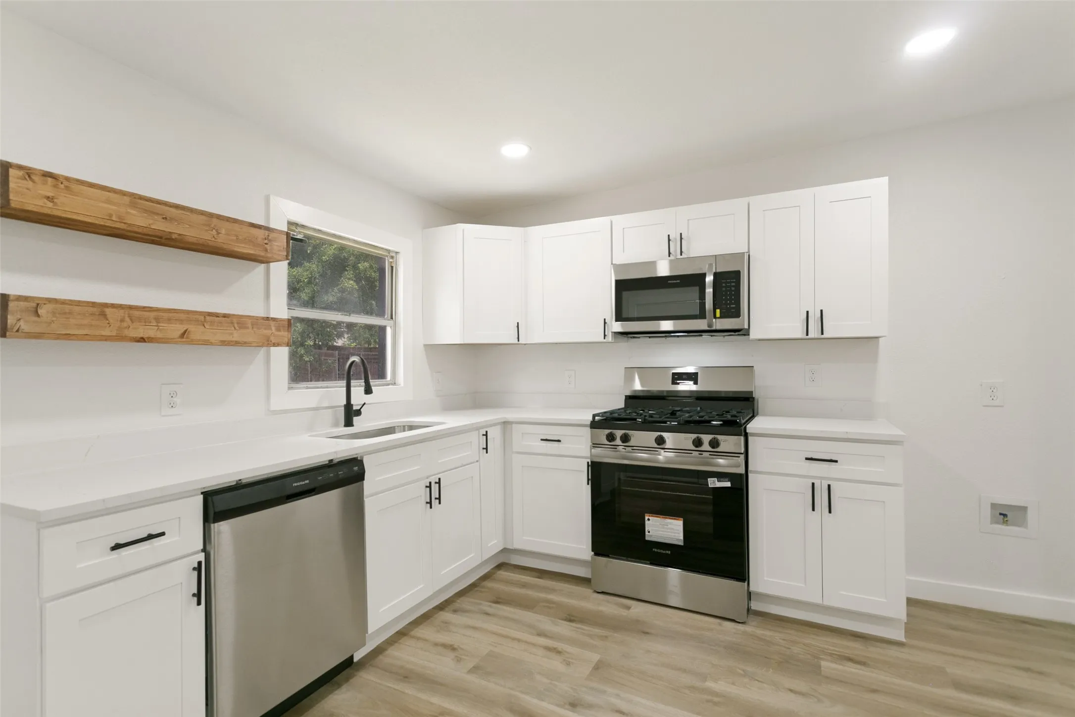 Modern kitchen featuring white cabinetry with matte black hardware, stainless steel appliances, light-toned countertops, and wood-finish flooring