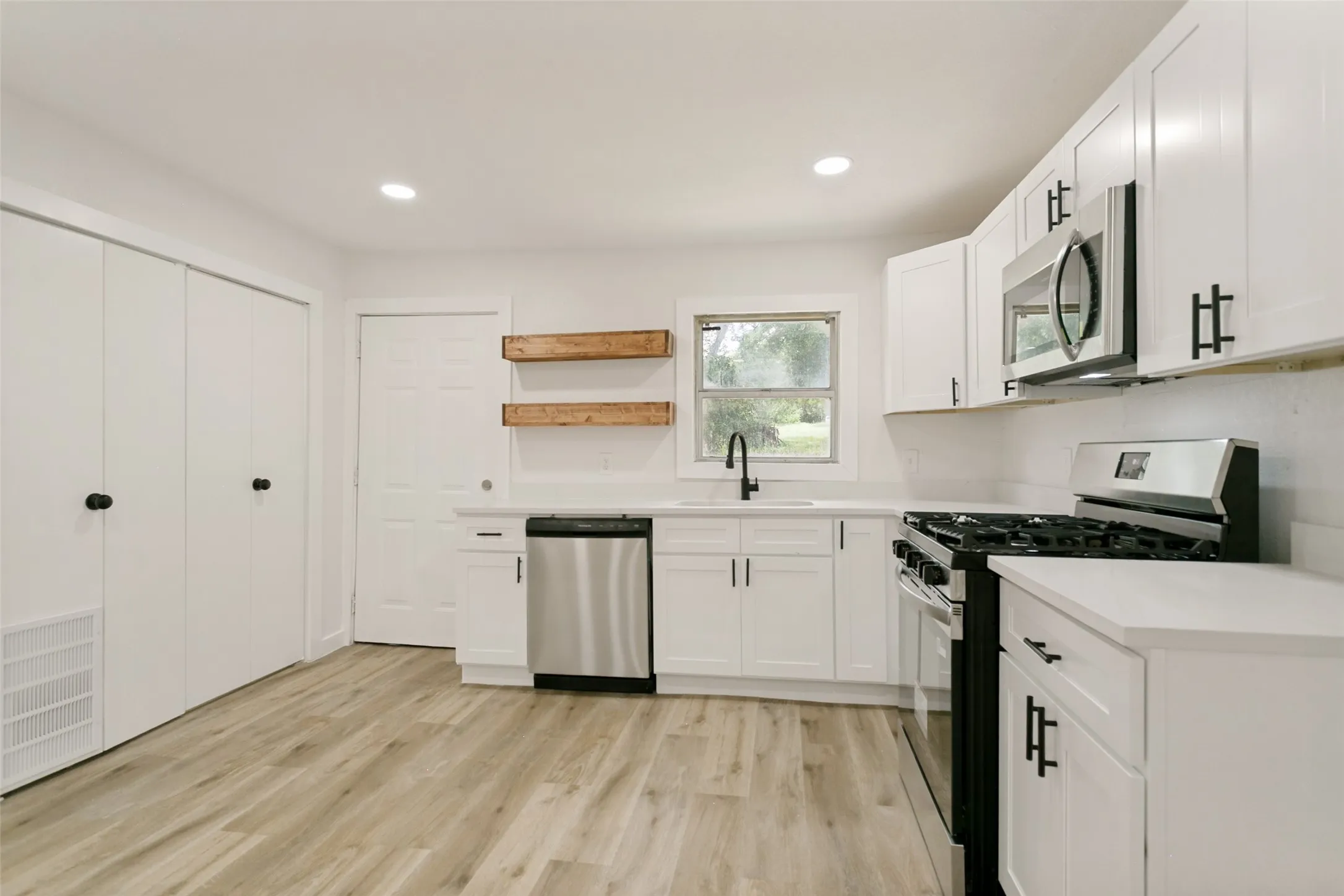Updated kitchen featuring white shaker cabinetry, stainless steel appliances, light-toned wood-finish flooring, and open shelving