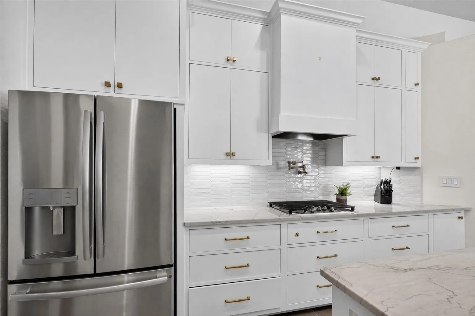 Kitchen featuring a stainless steel French-door refrigerator, white shaker cabinetry with gold-toned hardware, a gas cooktop, a white tile backsplash, and light-toned countertops