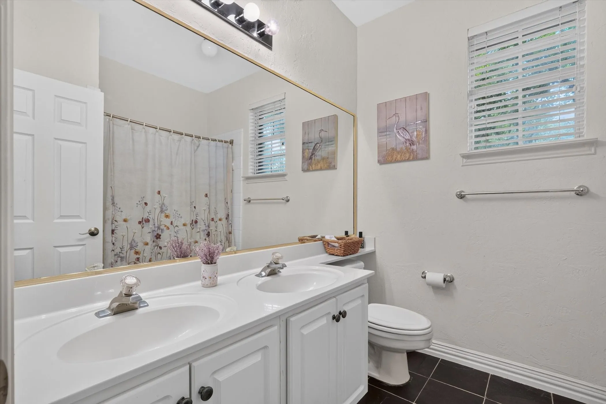 Bathroom featuring a dual-sink vanity with white cabinetry, black square floor tiling, a toilet, a window with blinds, and chrome fixtures